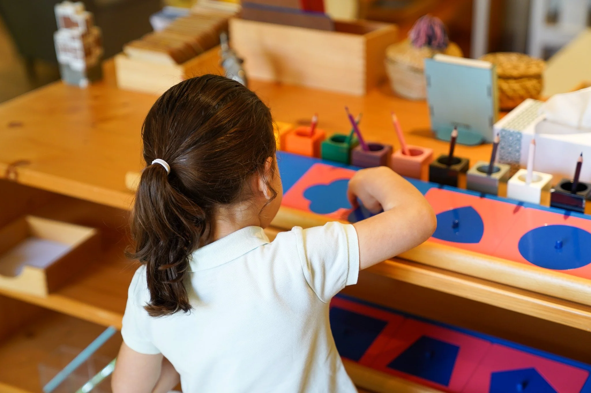 يدا طفل يعمل على رسم هندسي بقلم تحديد أحمر باستخدام مسطرة زرقاء مثلثة على ورقة بيضاء في لوحة رسم خشبية.