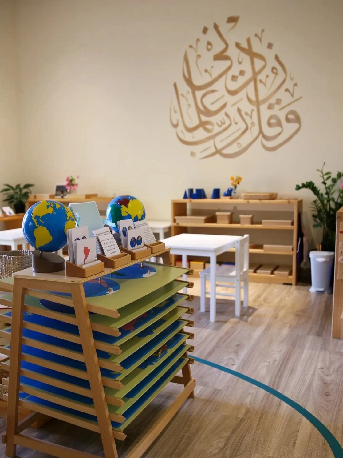 تصميم داخلي لفصل دراسي أو مساحة تعليمية تحتوي على مواد تعليمية بما في ذلك كرات أرضية وكتب وطاولة بيضاء مع كرسي. تظهر لوحات جدارية بالخط العربي ونباتات.