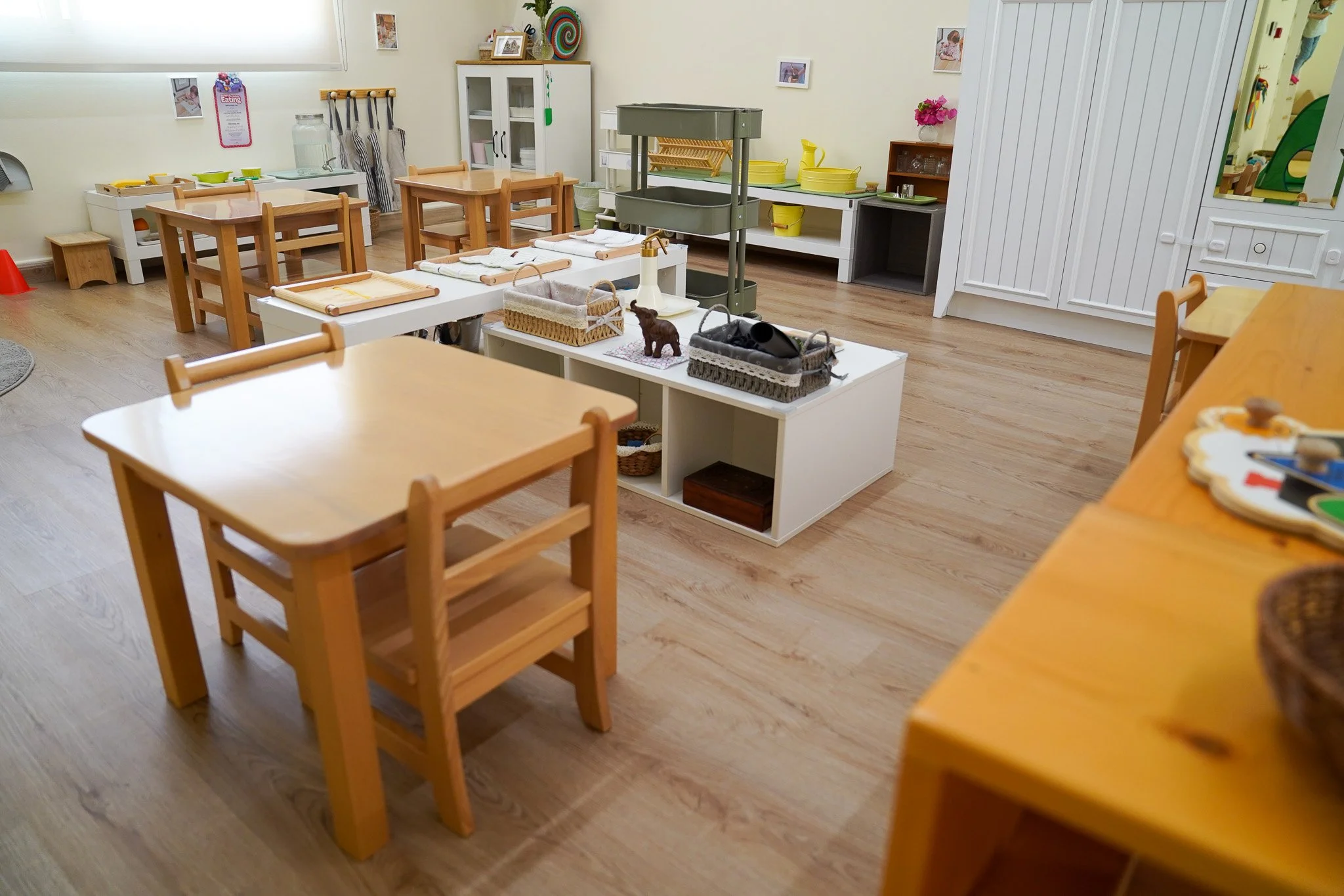 غرفة صف دراسي بها طاولات وكراسي خشبية صغيرة، وطاولة عرض بيضاء في المنتصف بها سلال وألعاب، ورفوف بها حاويات وزخارف ملونة. تحتوي الغرفة على أرضيات خشبية وجدران ذات ألوان فاتحة عليها صور.