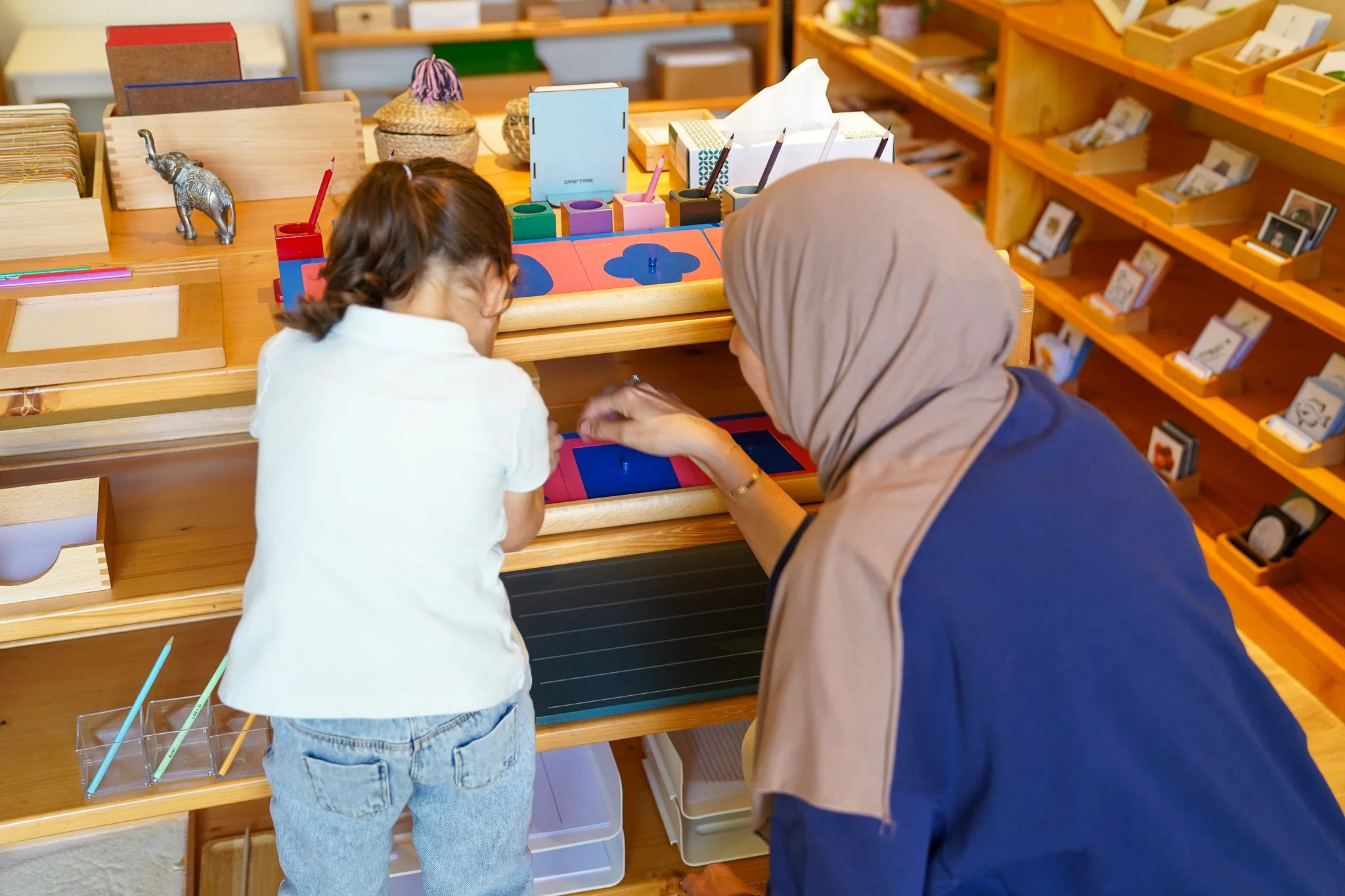 فتاة صغيرة وامرأة أكبر سناً، ربما تكون معلمة أو مقدمة رعاية، يشاركان في نشاط تعليمي على طاولة خشبية في فصل دراسي. ترتدي الفتاة قميصاً أبيض وبنطلون جينز أزرق، وترتدي المرأة حجاباً وسترة زرقاء. توجد أدوات تعليمية وزخرفية متنوعة على الرفوف والطاولة.