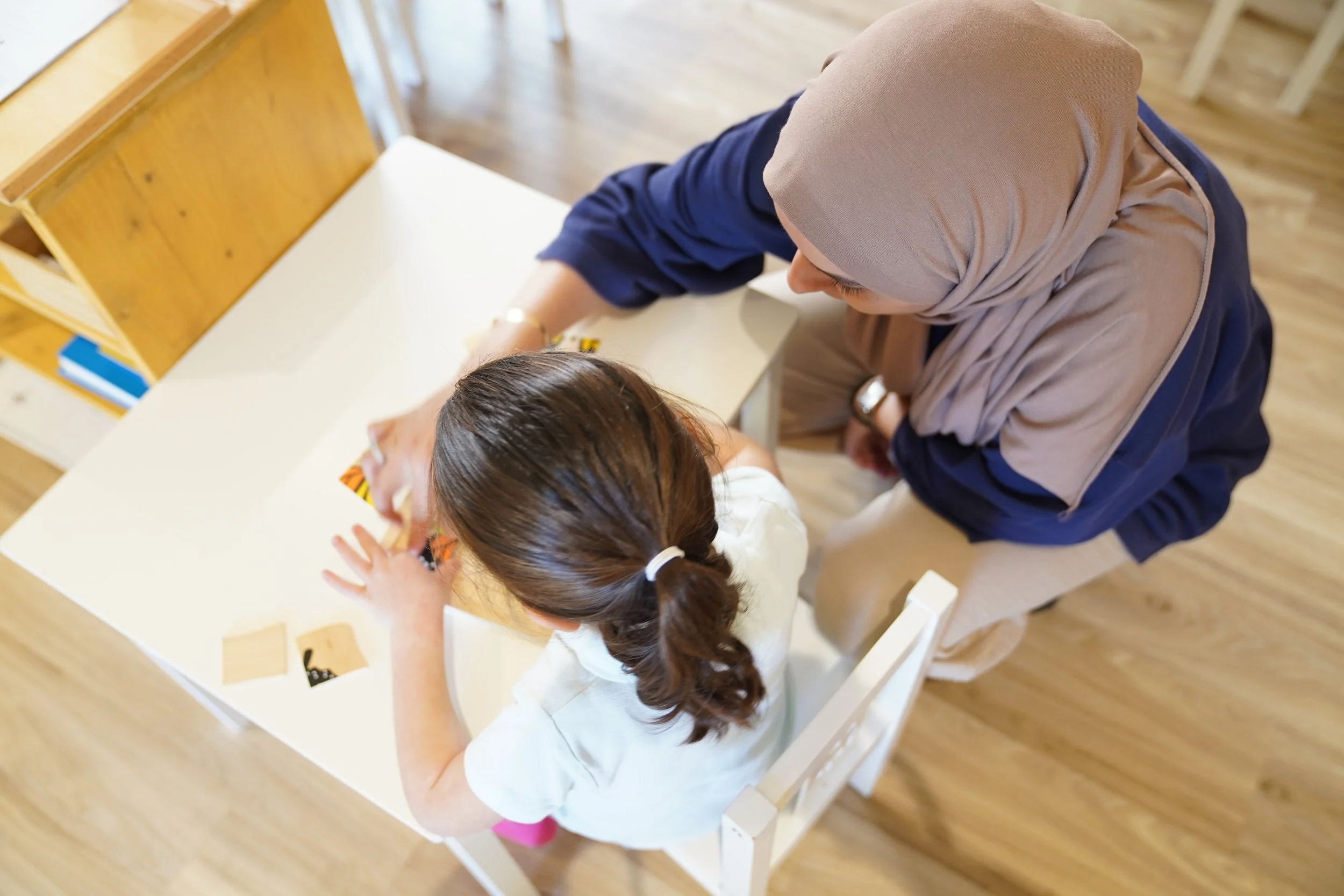 امرأة بالغة ترتدي حجاباً بيج اللون وتساعد فتاة صغيرة ذات شعر بني في ذيل حصان، على طاولة بيضاء ذات أرضية خشبية في الخلفية.