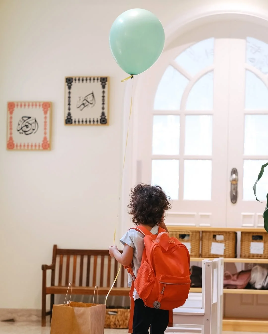 يقف طفل صغير بشعر مجعد يرتدي حقيبة ظهر ويحمل بالونًا أخضر في الداخل بالقرب من باب ذي نوافذ زجاجية بلورية وبجواره حقيبة تسوق ورقية ومقعدًا.