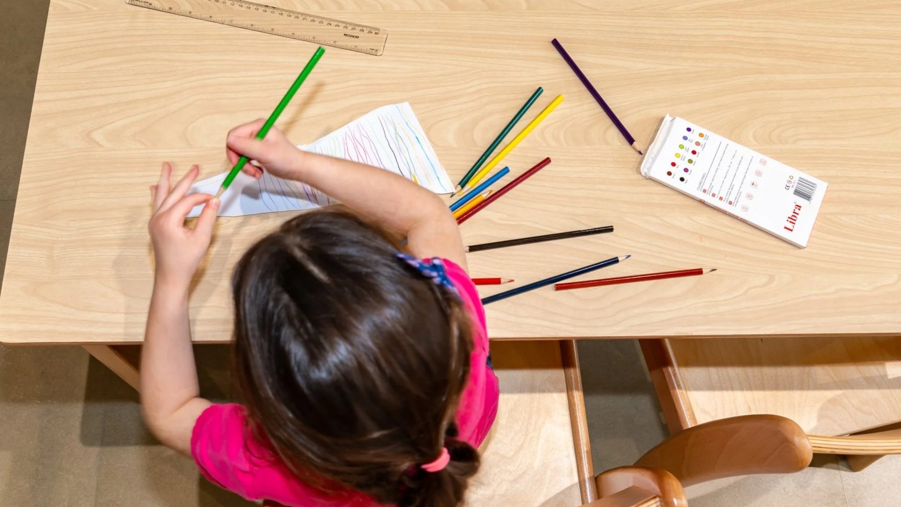 يدا طفل يعمل على رسم هندسي بقلم تحديد أحمر باستخدام مسطرة زرقاء مثلثة على ورقة بيضاء في لوحة رسم خشبية.