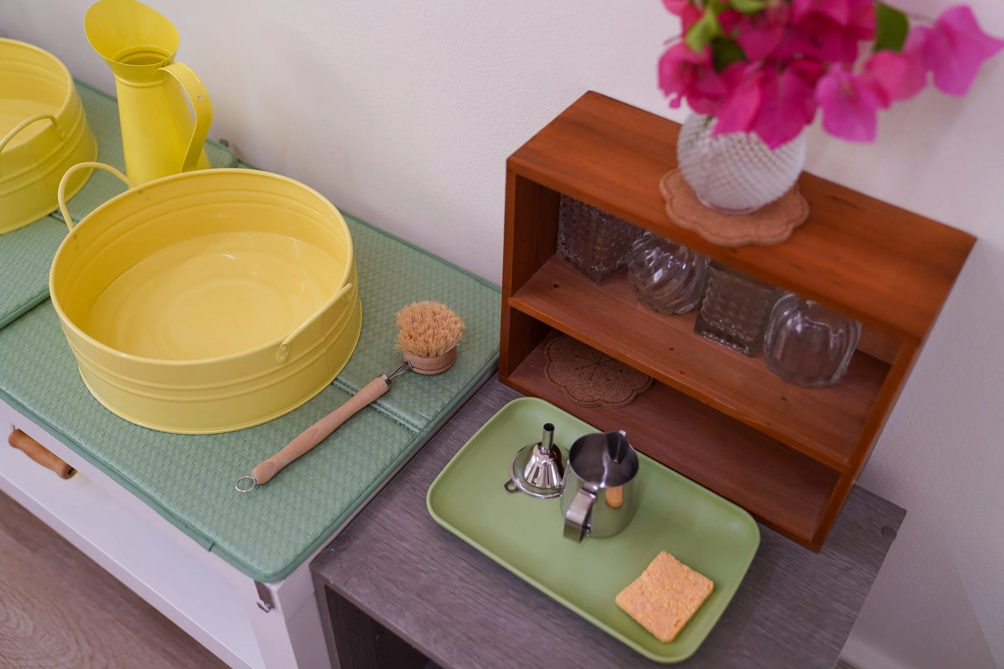 مجموعة من أدوات التنظيف والتنظيم المنزلية على طاولة صغيرة. توجد مغاسل بلاستيكية صفراء وممسحة صفراء صغيرة وإسفنجة خضراء وصينية خضراء مع غلاية فضية وقمع صغير وكوب أسود. يوجد رف خشبي بالأعلى يحمل مزهريات زجاجية ومزهرية بيضاء بها زهور وردية.