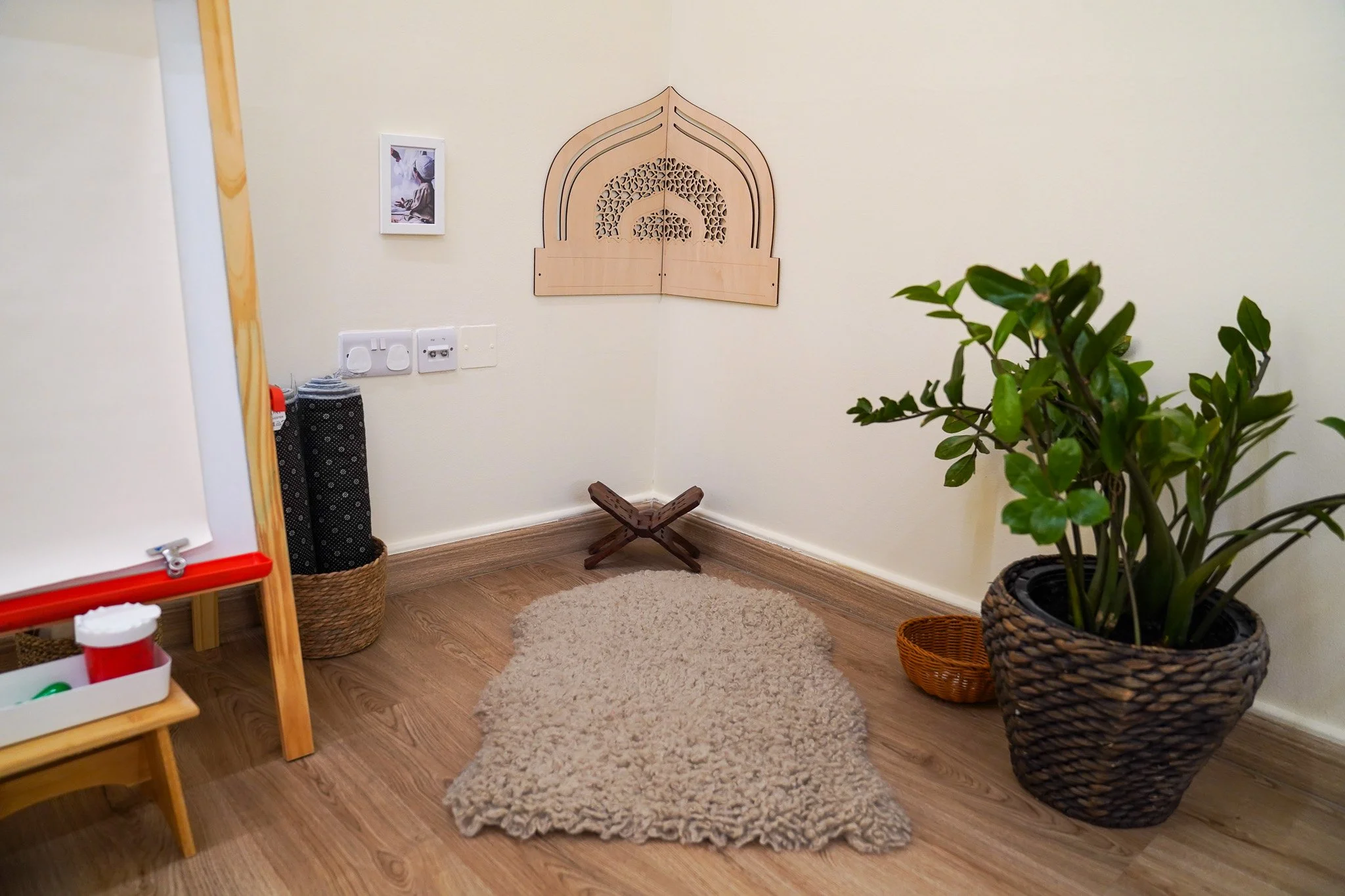 زاوية غرفة بها حائط بيج، وسجادة صغيرة بيج، ونبتة كبيرة بوعاء كبير، وسلة منسوجة، وزخرفة حائط خشبية، وصورة فوتوغرافية صغيرة.