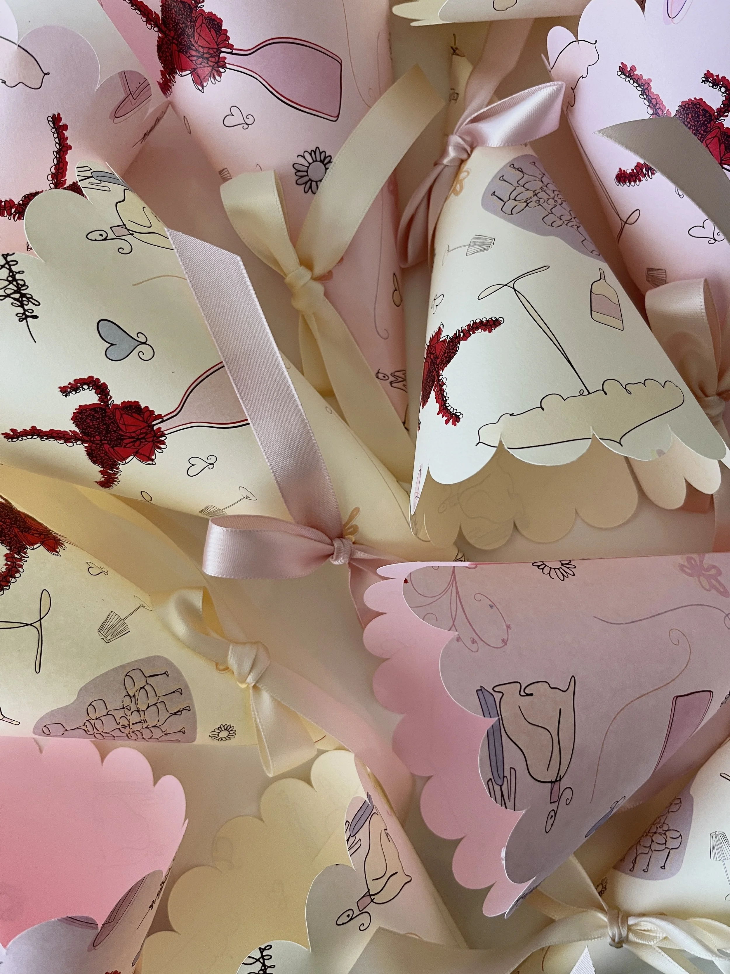 Pink and yellow confetti cones decorated with illustrations of cakes, teapots, and hearts, tied with ribbons, arranged in a pile.