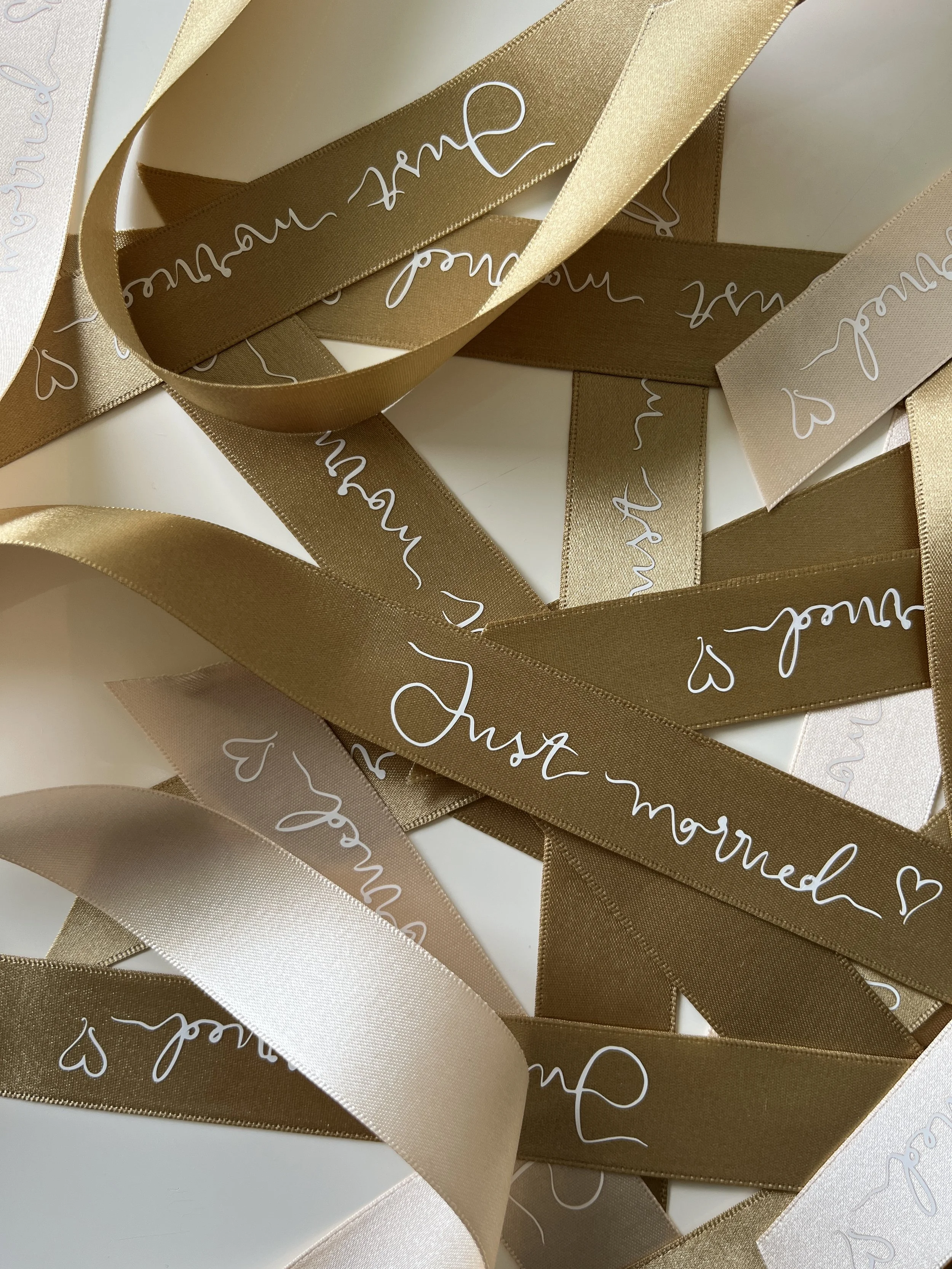 Multiple strips of metallic gold, beige, and cream-colored ribbons with the phrase 'Just married' and small hearts written in white cursive on them, spread out in a random pattern.