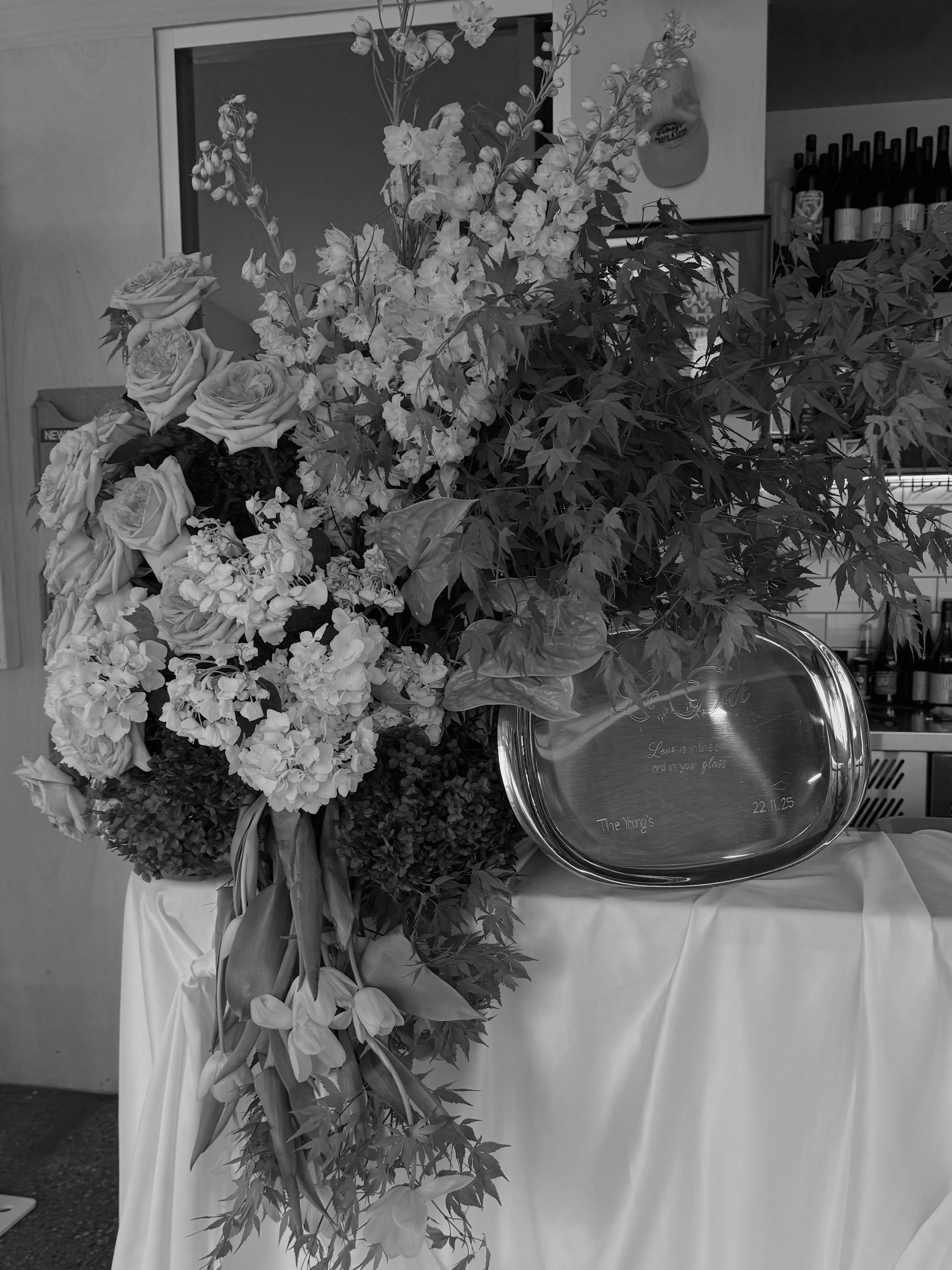 A large floral arrangement with roses, hydrangeas, and other flowers on a table covered with a cloth, with a silver tray reflecting engraved text and a date, in a setting with bottles and a mirror in the background.