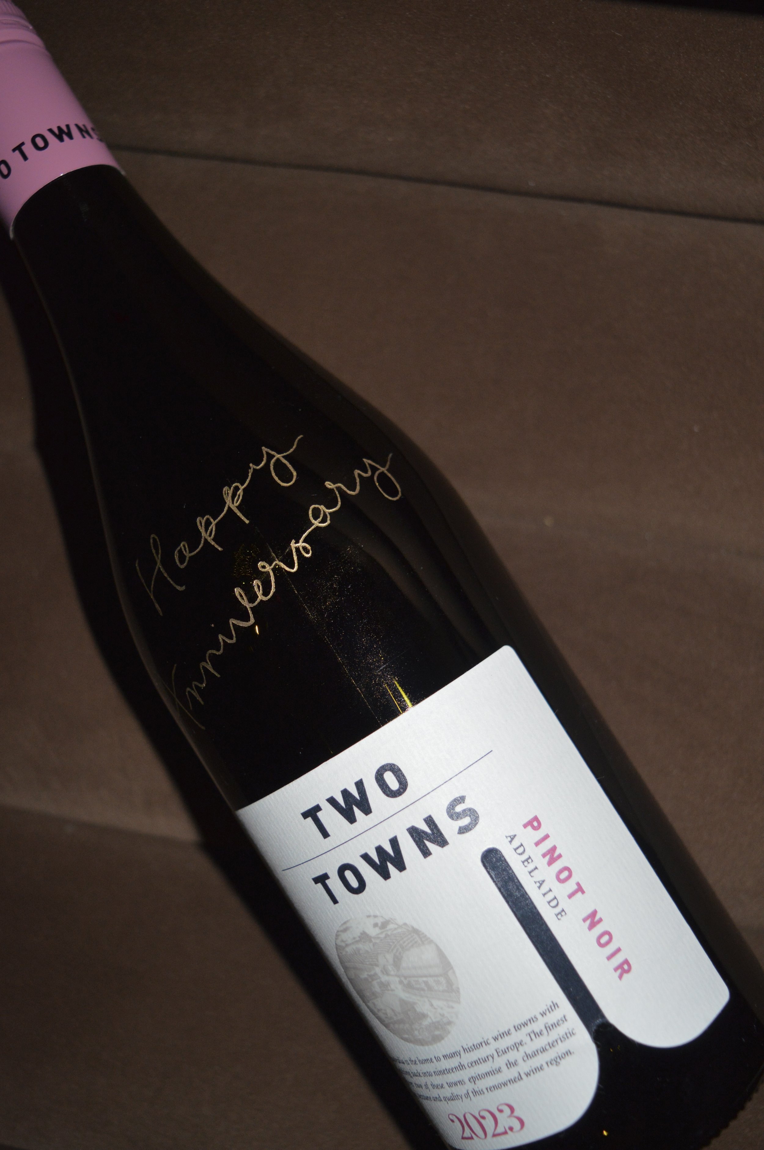 A wine bottle lying on a brown surface with a white label reading "Two Towns Pinot Noir Adelaide 2023" and gold script writing that says "happening memories."