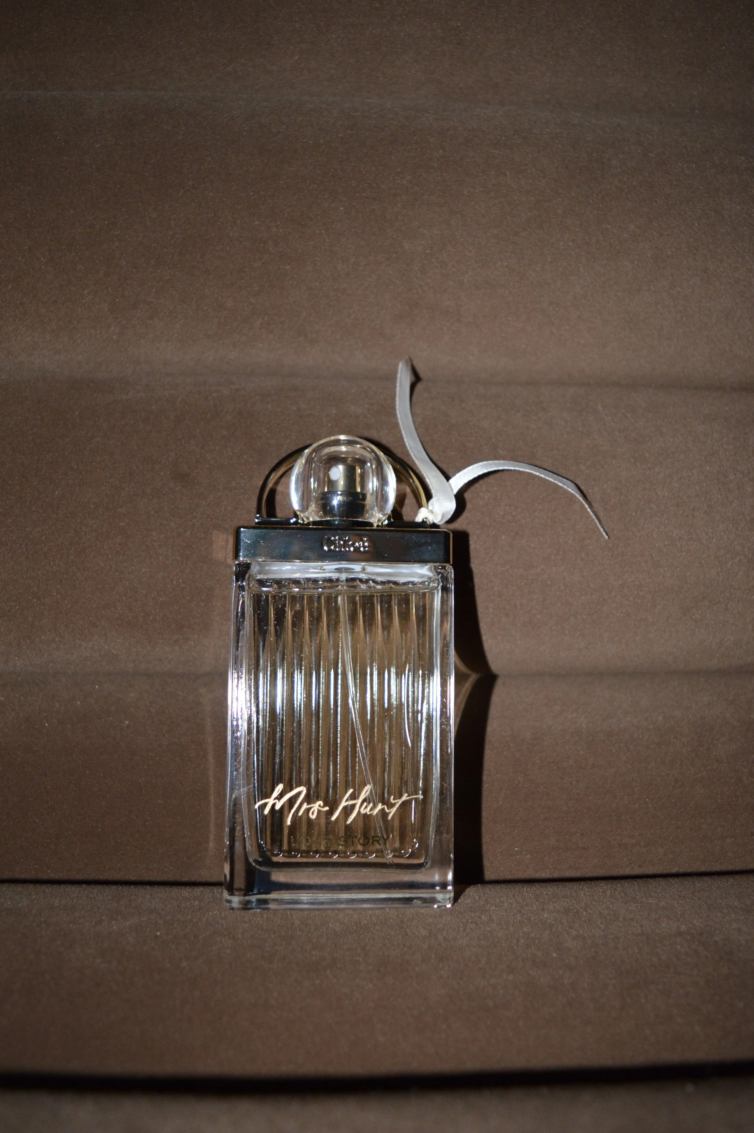 A clear glass perfume bottle with a metal cap and a decorative silver leaf attached, labeled 'Mrs. Harty', placed on a brown fabric background.