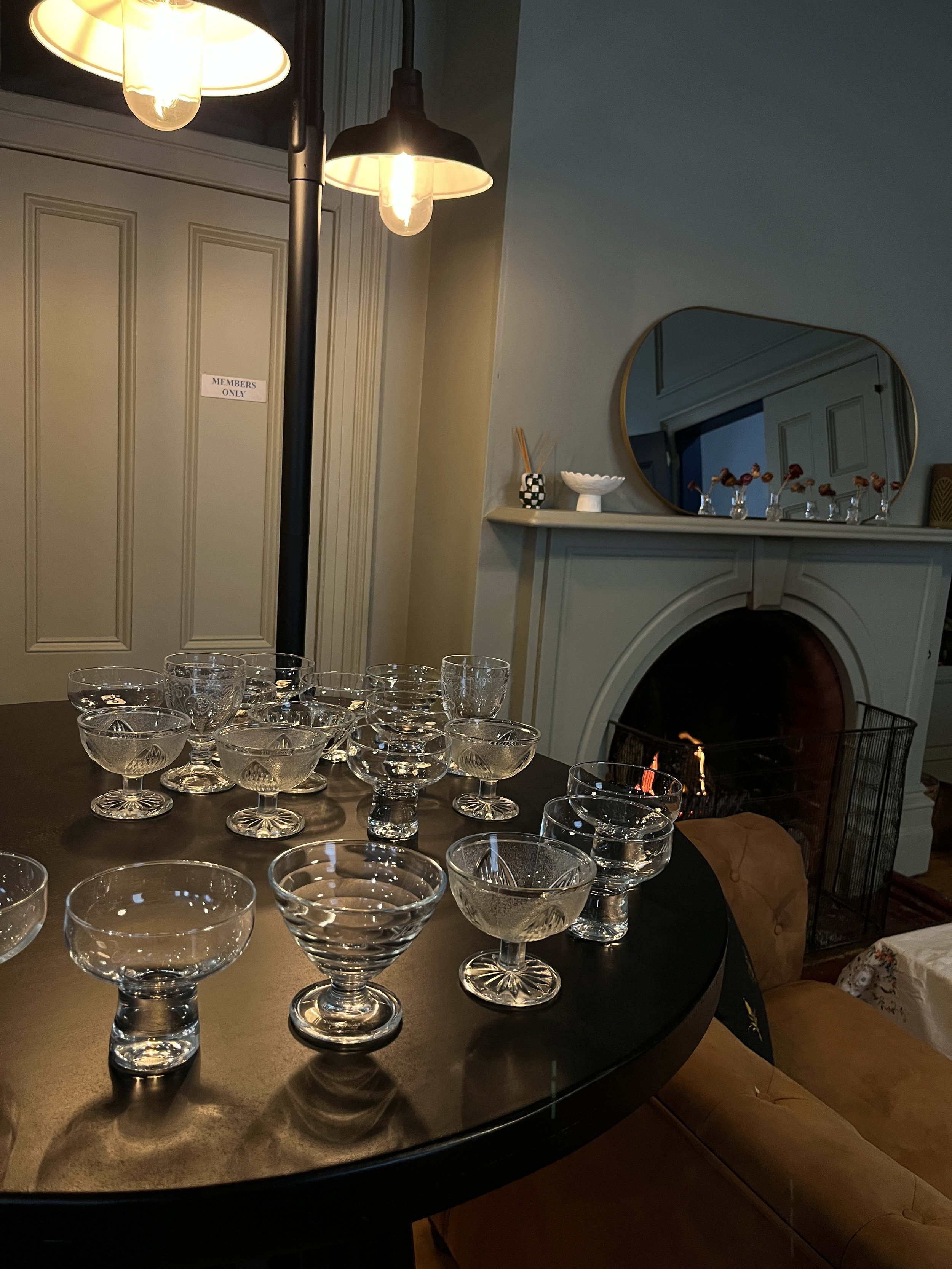 A dining room with a round table set with multiple glass dessert cups. There is a cozy fireplace with a mirror above it, decorative small bottles with flowers, and a tall floor lamp with four exposed light bulbs. A sign on the door reads 'MEMBERS ONLY'.