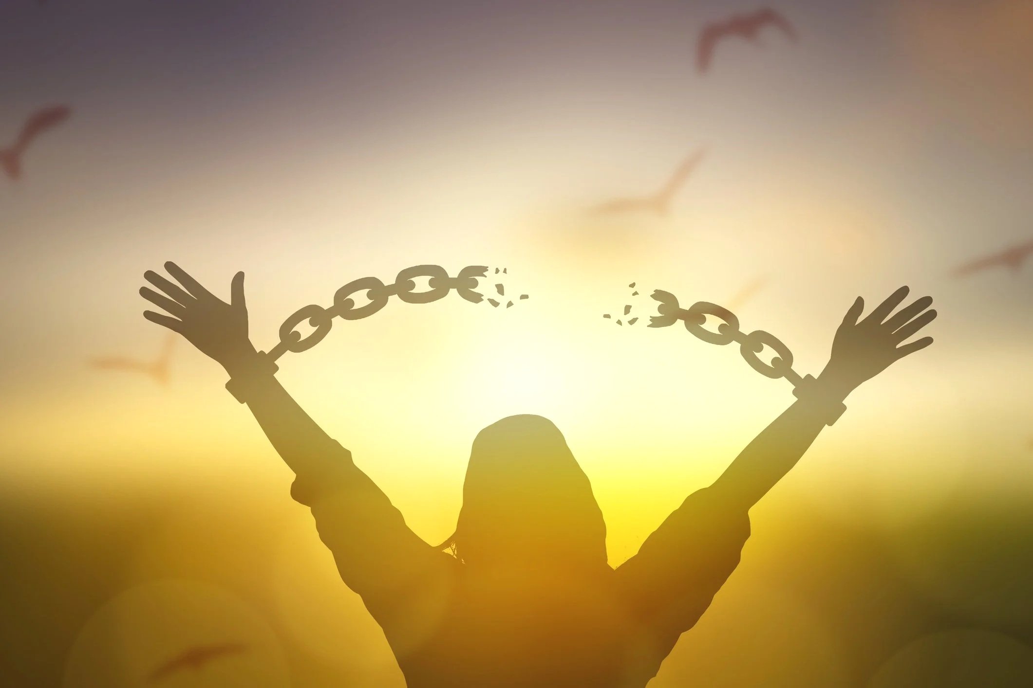 Silhouette of a person with hands raised in front of a sunset or sunrise sky, with broken chains around their wrists and hands, symbolizing freedom or breaking free.