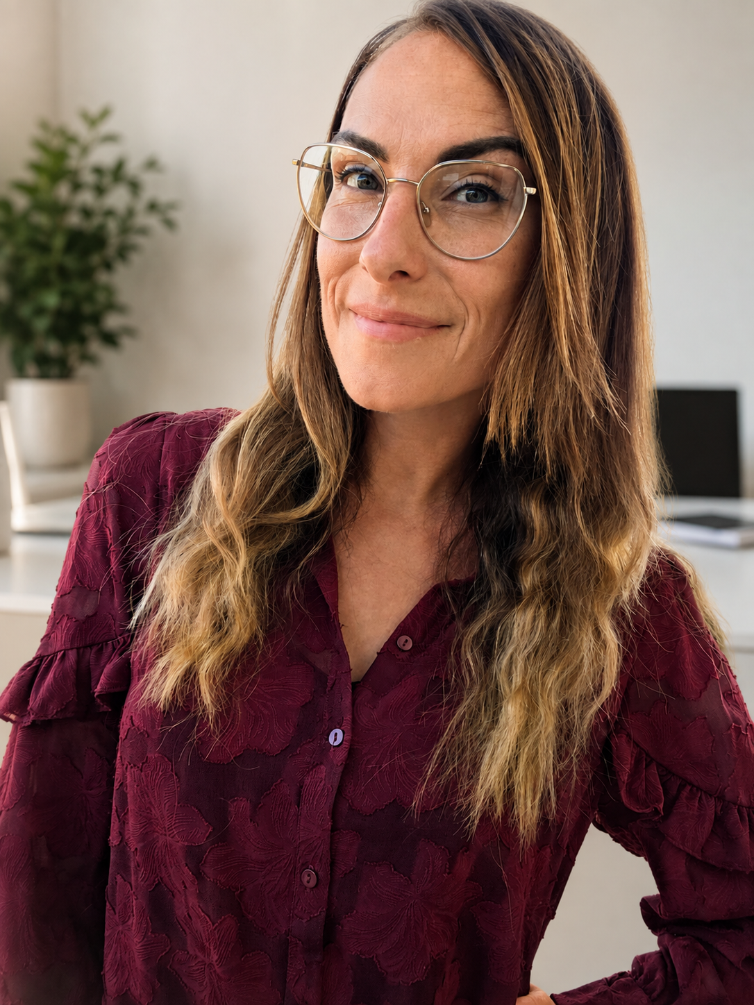 A woman with long wavy hair wearing glasses and a burgundy patterned blouse, smiling slightly, seated in an office with a blurred background that includes a potted plant and a monitor. Home office, virtual assistant, administration