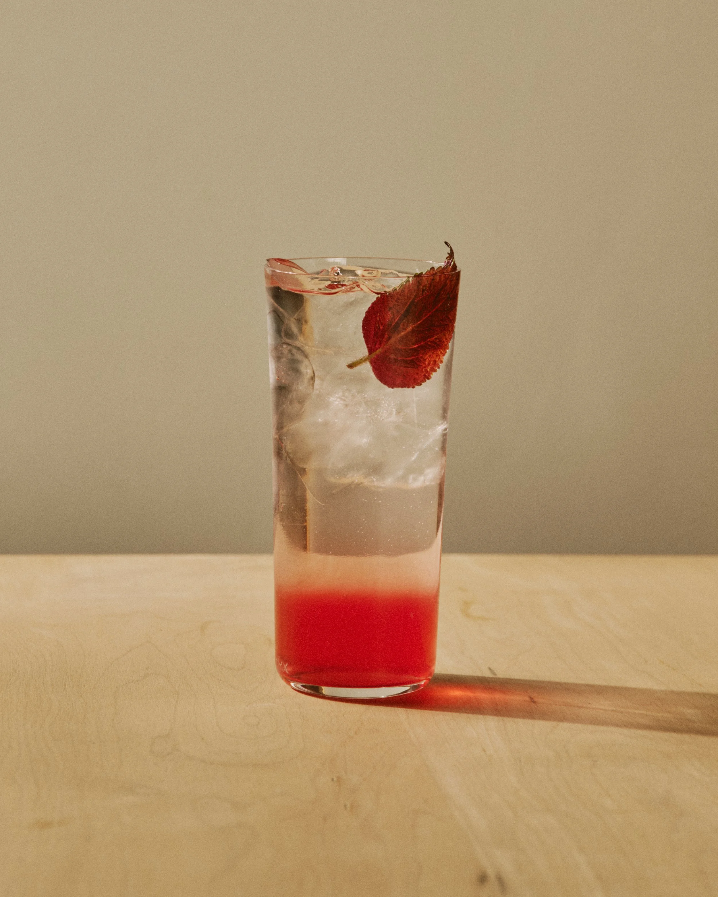 A tall glass filled with a layered pink and clear beverage, ice cubes, and red leaves, sitting on a wooden surface with a plain light background.