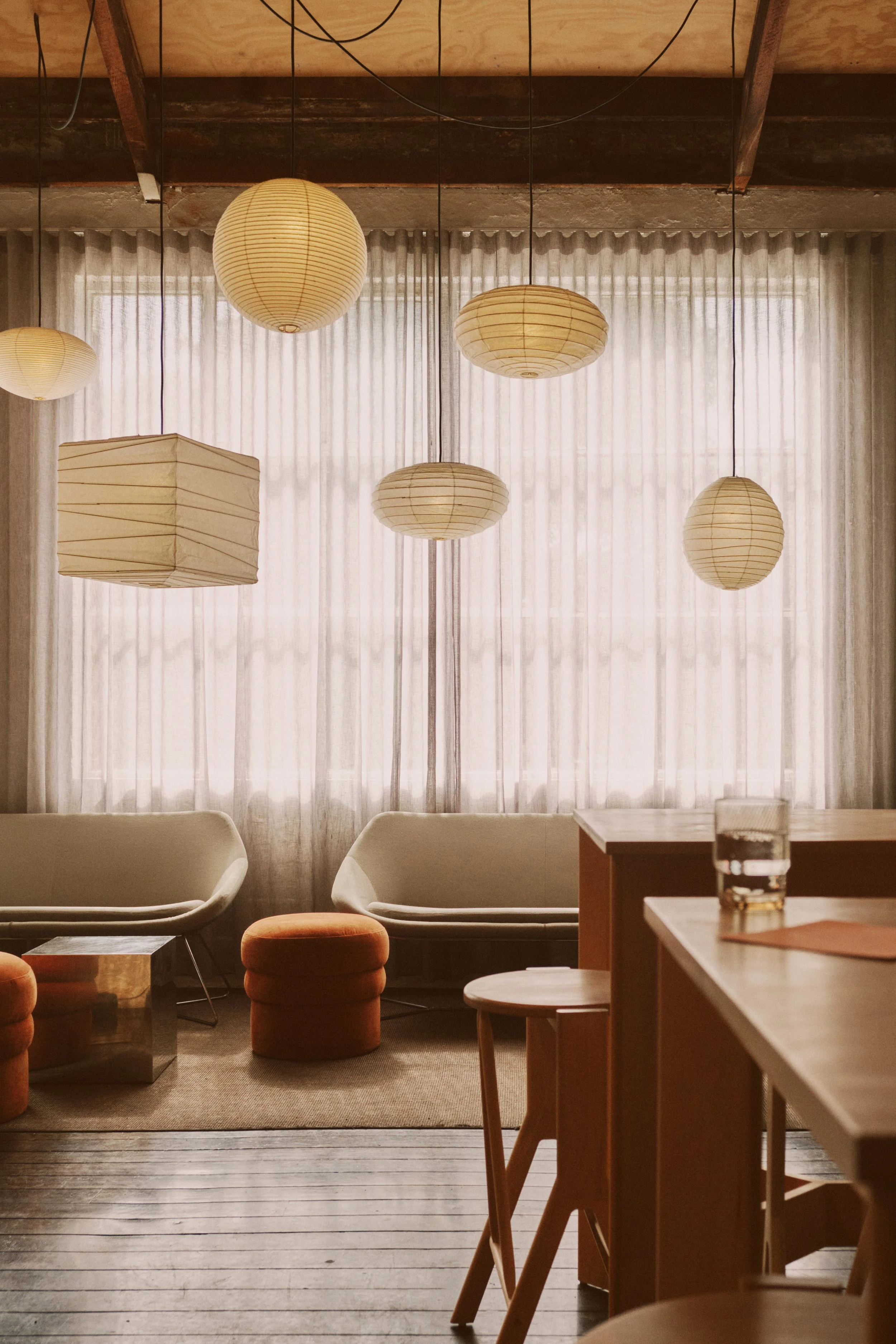Interior of a cozy lounge with hanging paper lanterns, two beige sofas, and small stools near large windows with sheer curtains, illuminated by natural light.