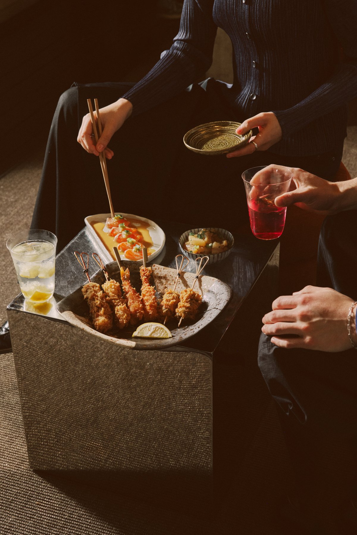A table with various Japanese dishes including kushi katsu, a small plate of sashimi, a small bowl of pickled vegetables, and drinks, with people serving and enjoying the meal.