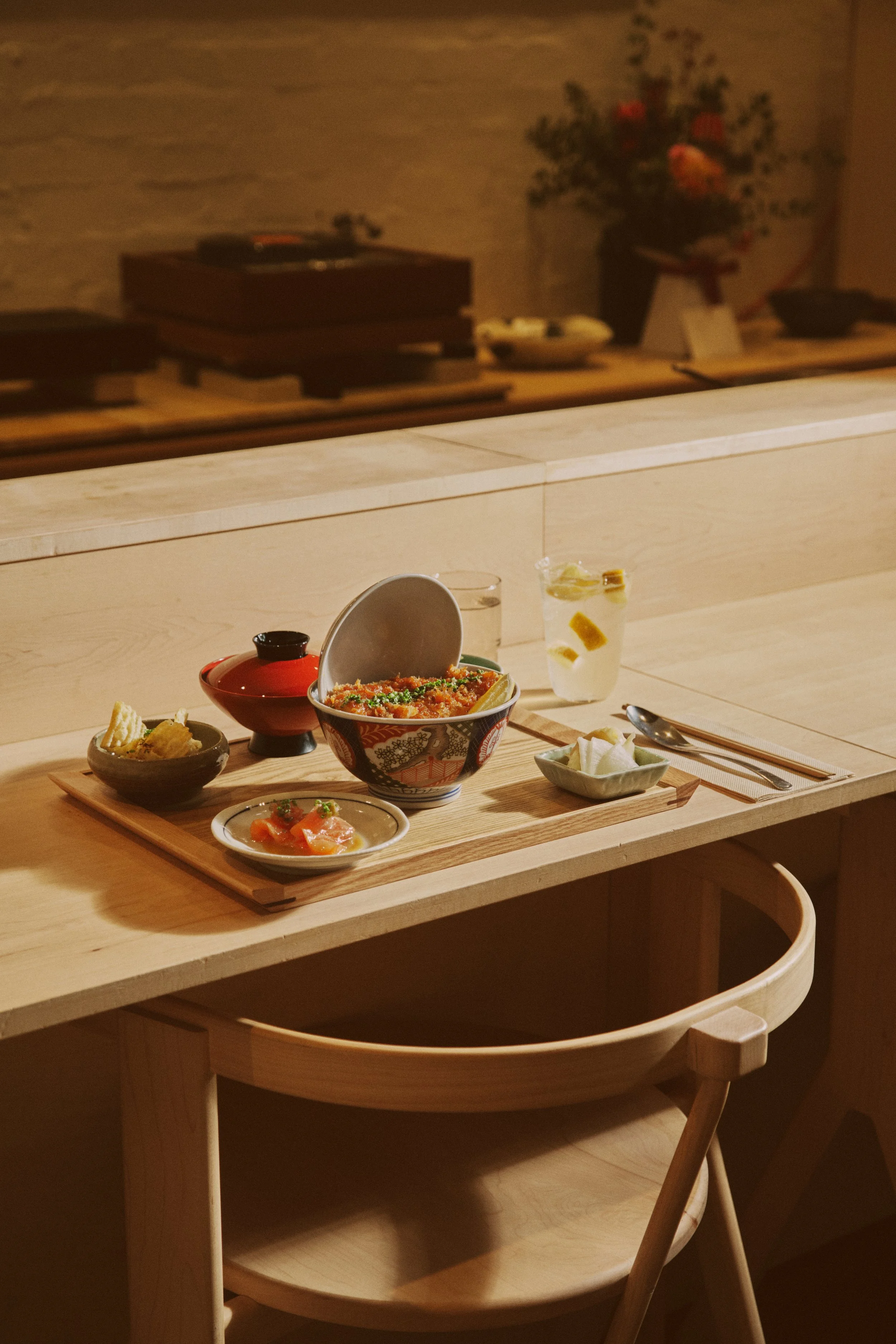 A katsudon meal set on a wooden tray including a bowl of rice topped with vegetables or seafood, a small dish of sashimi, a side dish, and a glass of lemon sour, placed on a wooden counter in a softly lit restaurant.