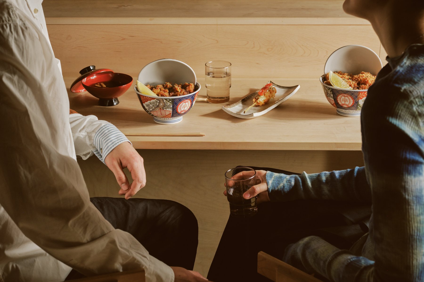 Two people sitting at a wooden table with bowls of food and a glass of water between them. The table has a kushi katsu and two bowls of katsu don.