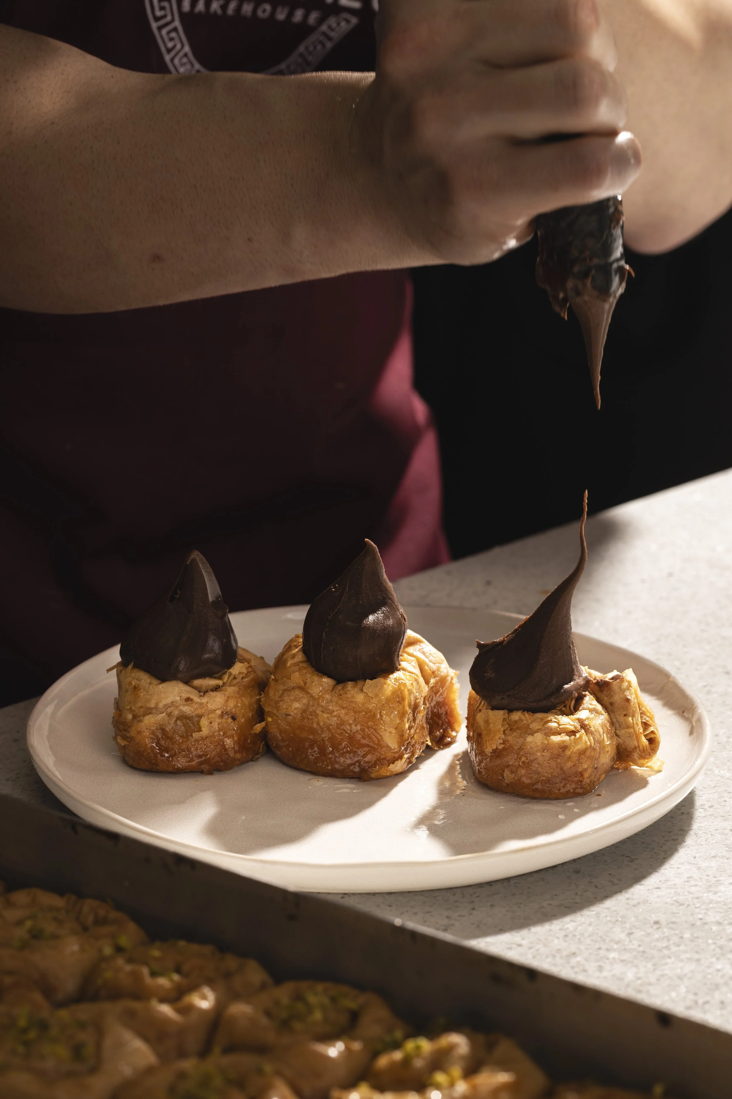 A person decorating dessert pastries with chocolate on a white plate, with another plate of similar pastries in the foreground.
