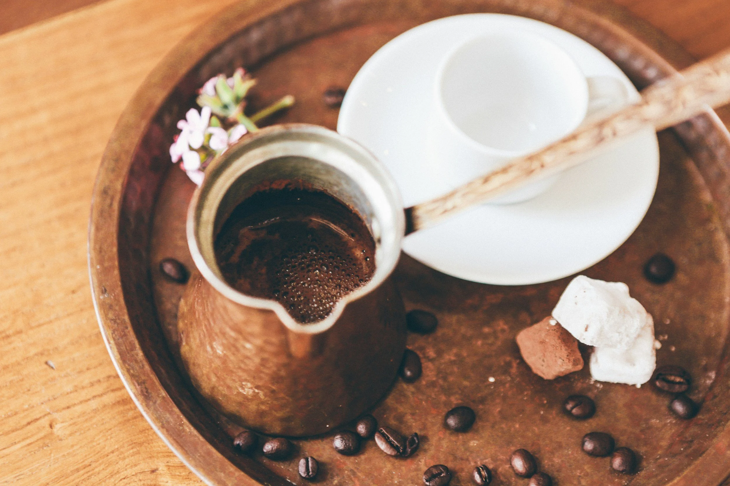 A coffee mug with black coffee, a small white creamer pitcher, a broken cookie, and scattered coffee beans on a wooden tray.