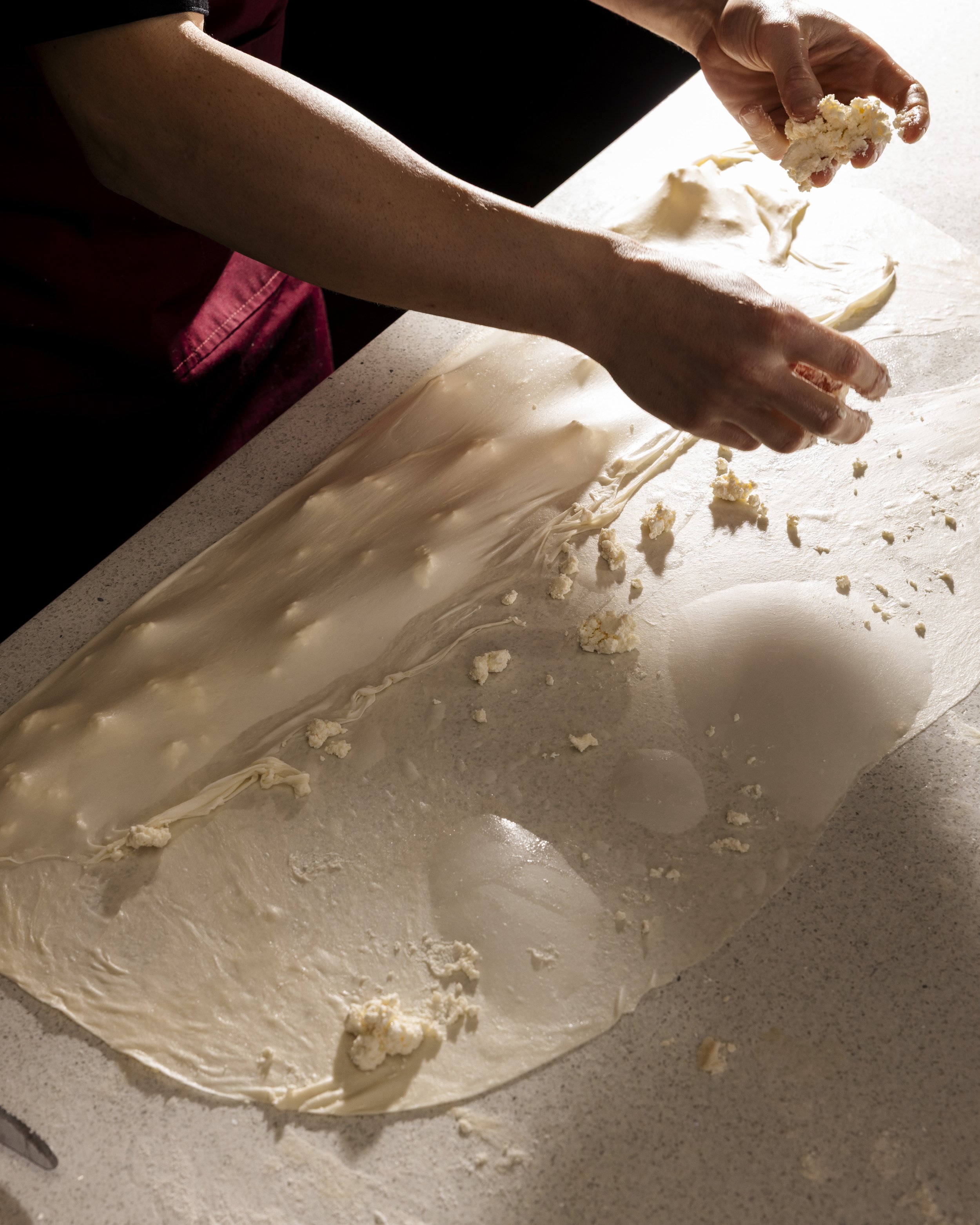 A person preparing pastry dough on a countertop. They are adding crumbled cheese or filling onto the dough, which is rolled out flat. Tiropita. greek pastries.