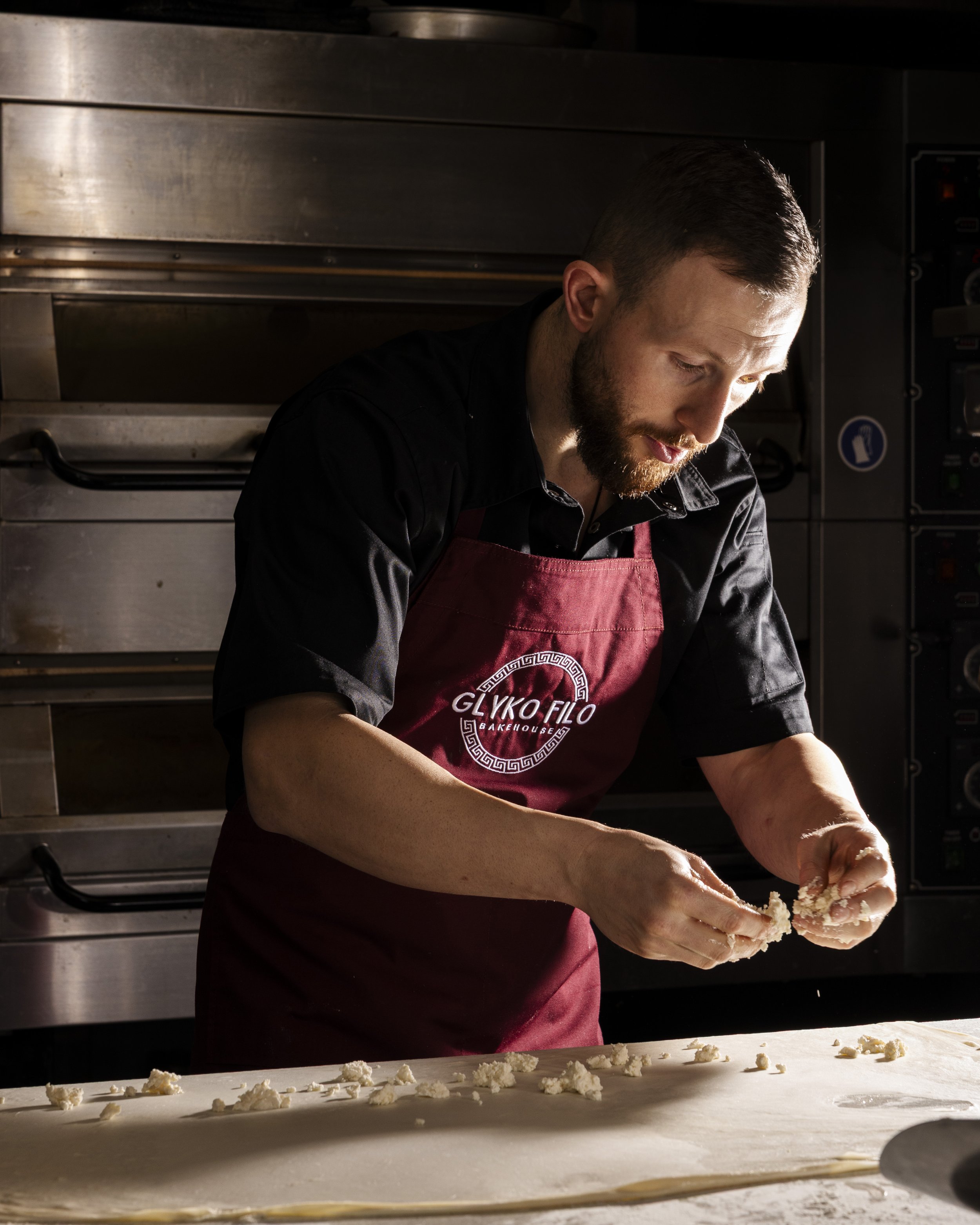 A man wearing a black shirt and a red apron with the logo 'Glyko Filo Bakehouse' prepares dough on a work surface in a professional kitchen, with stainless steel oven in the background.