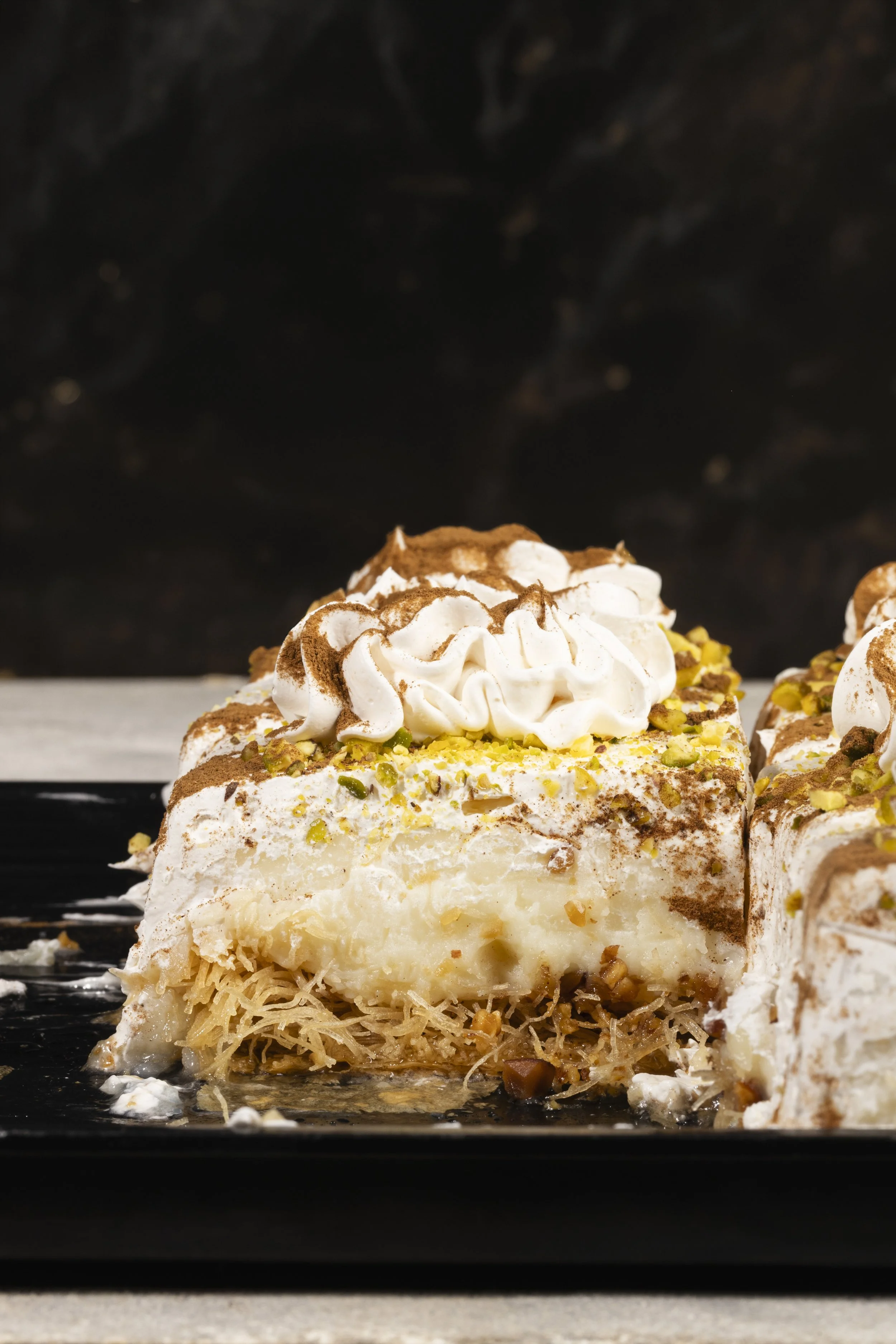 A slice of baklava topped with whipped cream, chopped pistachios, and cocoa powder on a black plate.