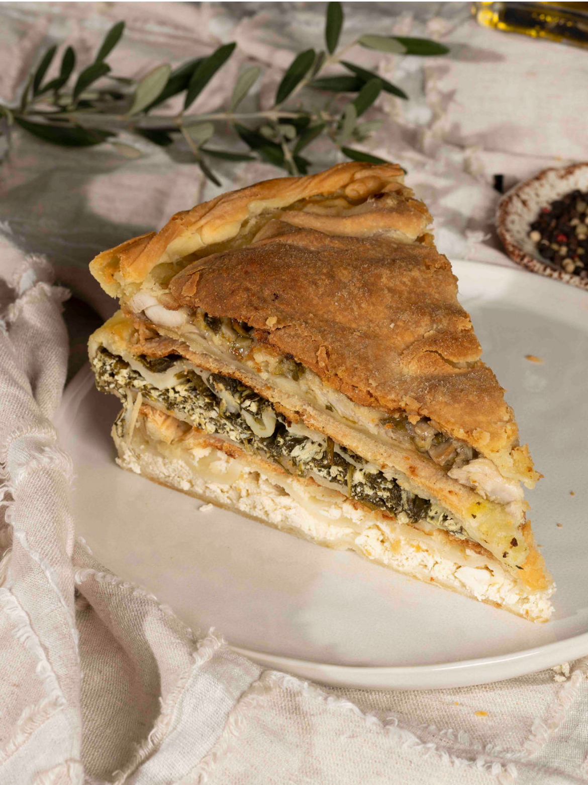 A slice of layered Spanakopita with a golden brown crust on a white plate, with herbs and vegetables inside, placed on a beige linen cloth with some leaves and a small bowl of pepper in the background. Traditional greek pastries.