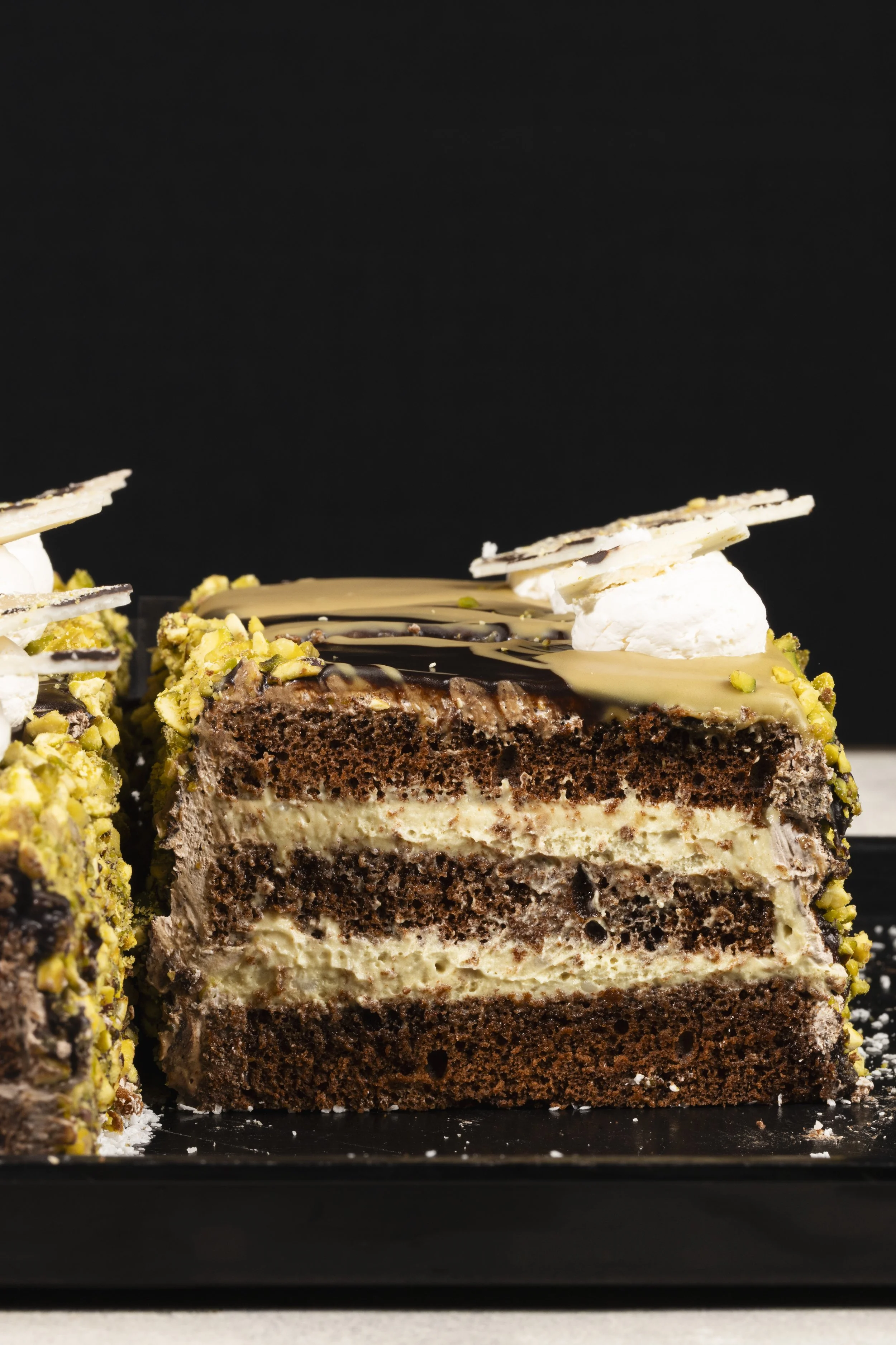 Slices of layered chocolate and vanilla cake with pistachios, chocolate glaze, and white chocolate decorations on a black background.