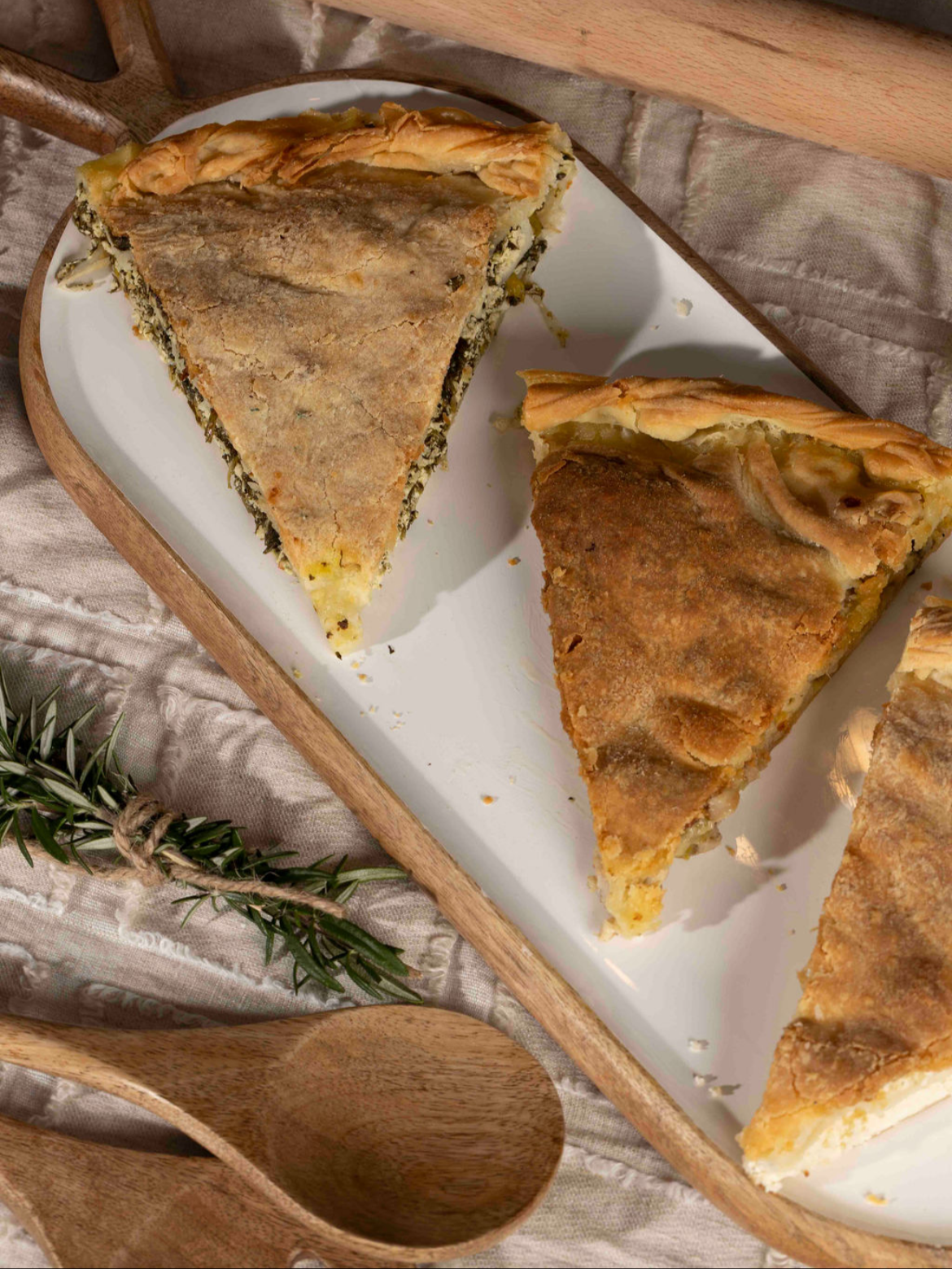 Slices of savory pie with golden crust on a white rectangular plate, with a wooden spoon and sprig of rosemary nearby.