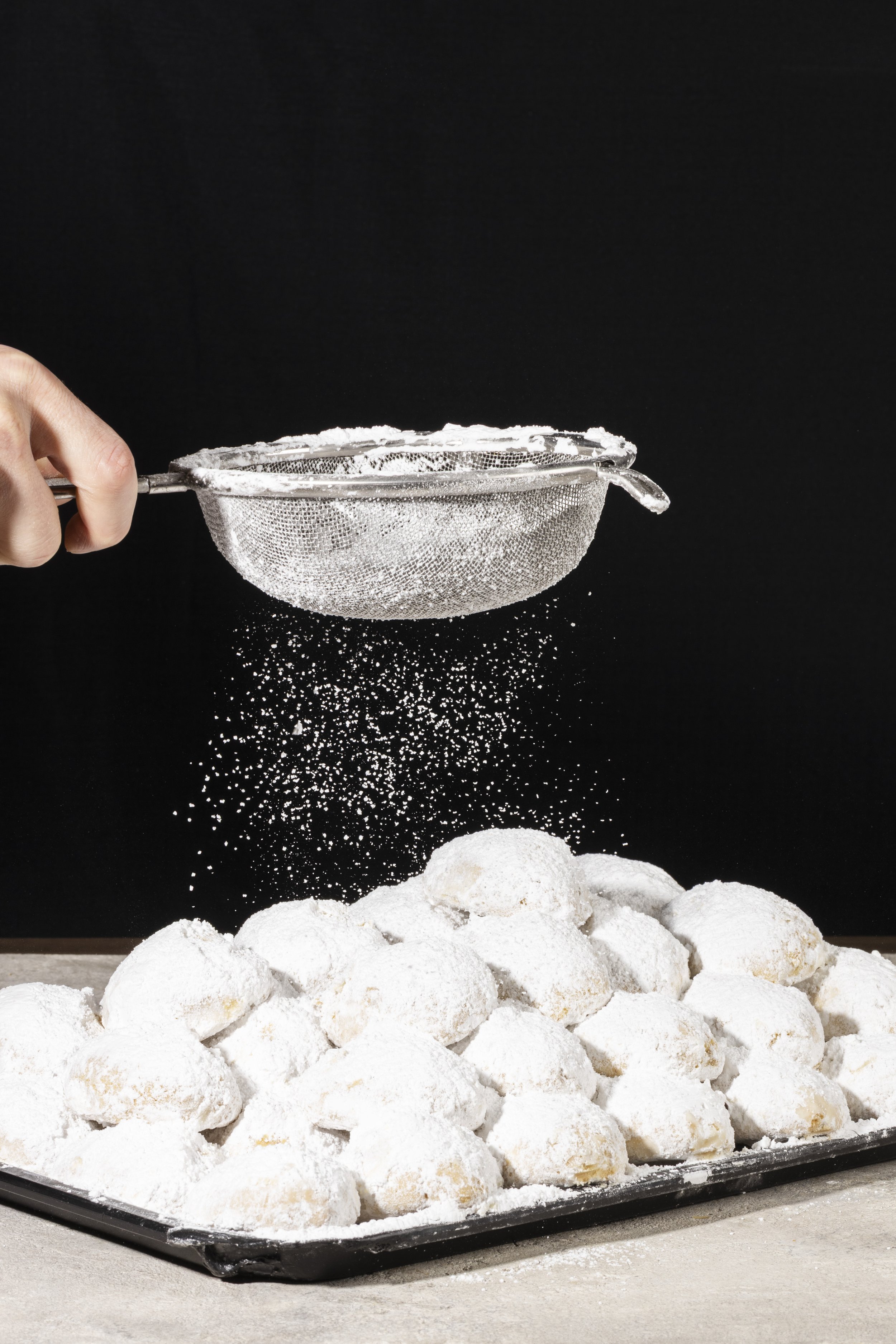 A person sifting powdered sugar over a pile of cookie dough balls on a black tray, with some sugar falling into the dough.
