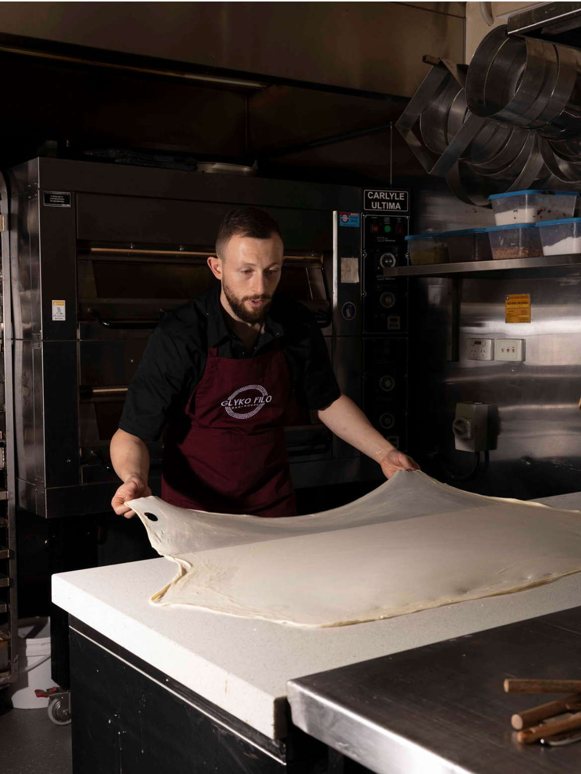 A man in a black shirt and maroon apron is stretching a large sheet of dough on a white countertop in a commercial kitchen.