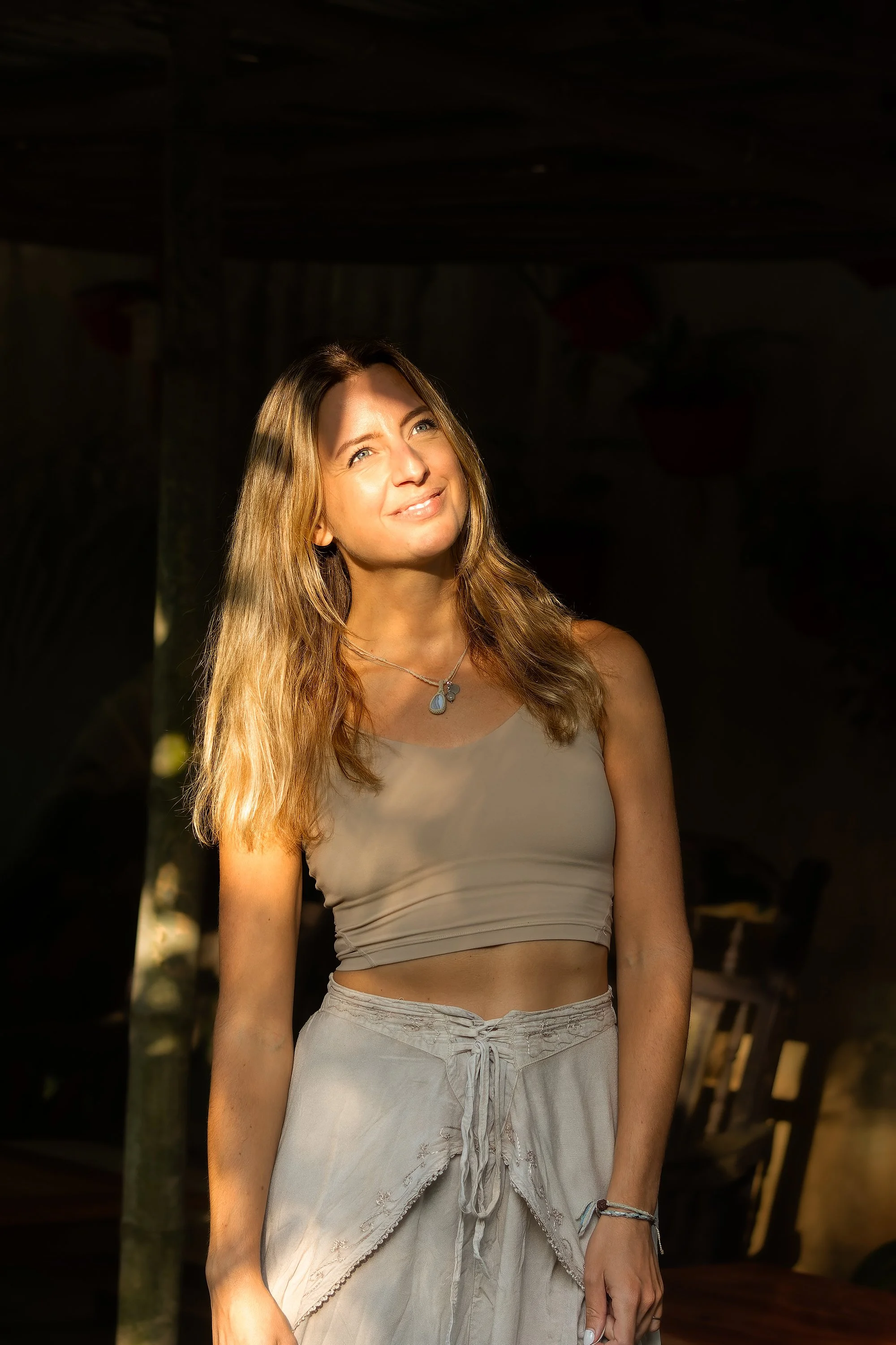 A woman with long, wavy, light brown hair is smiling and looking slightly upward. She is wearing a beige sleeveless top, light-colored pants with a drawstring, a necklace, and a few bracelets. The background appears to be a dark, covered outdoor area with some plants.