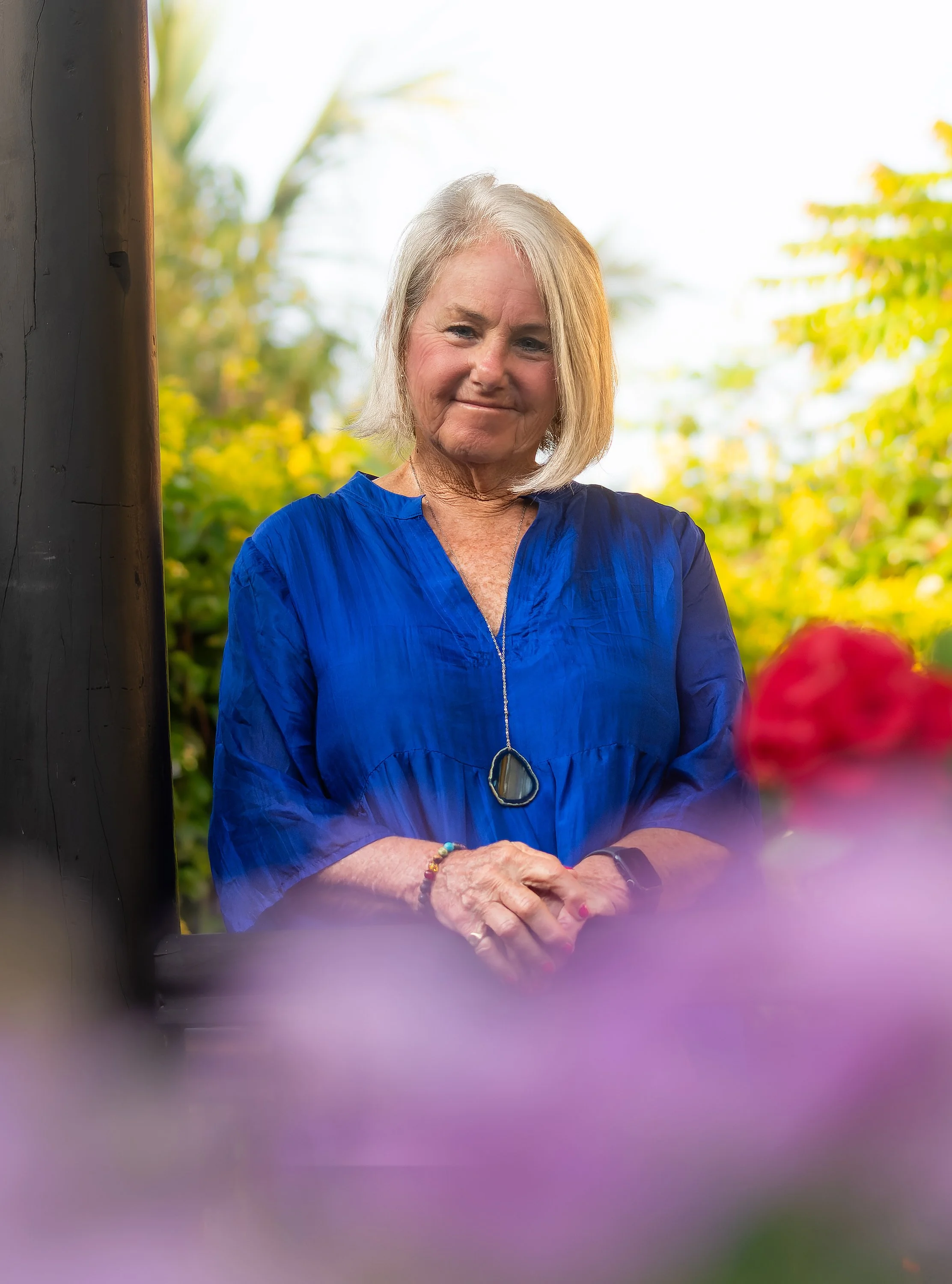 An older woman with gray hair smiling, wearing a blue blouse and necklace, standing outdoors with greenery in the background.