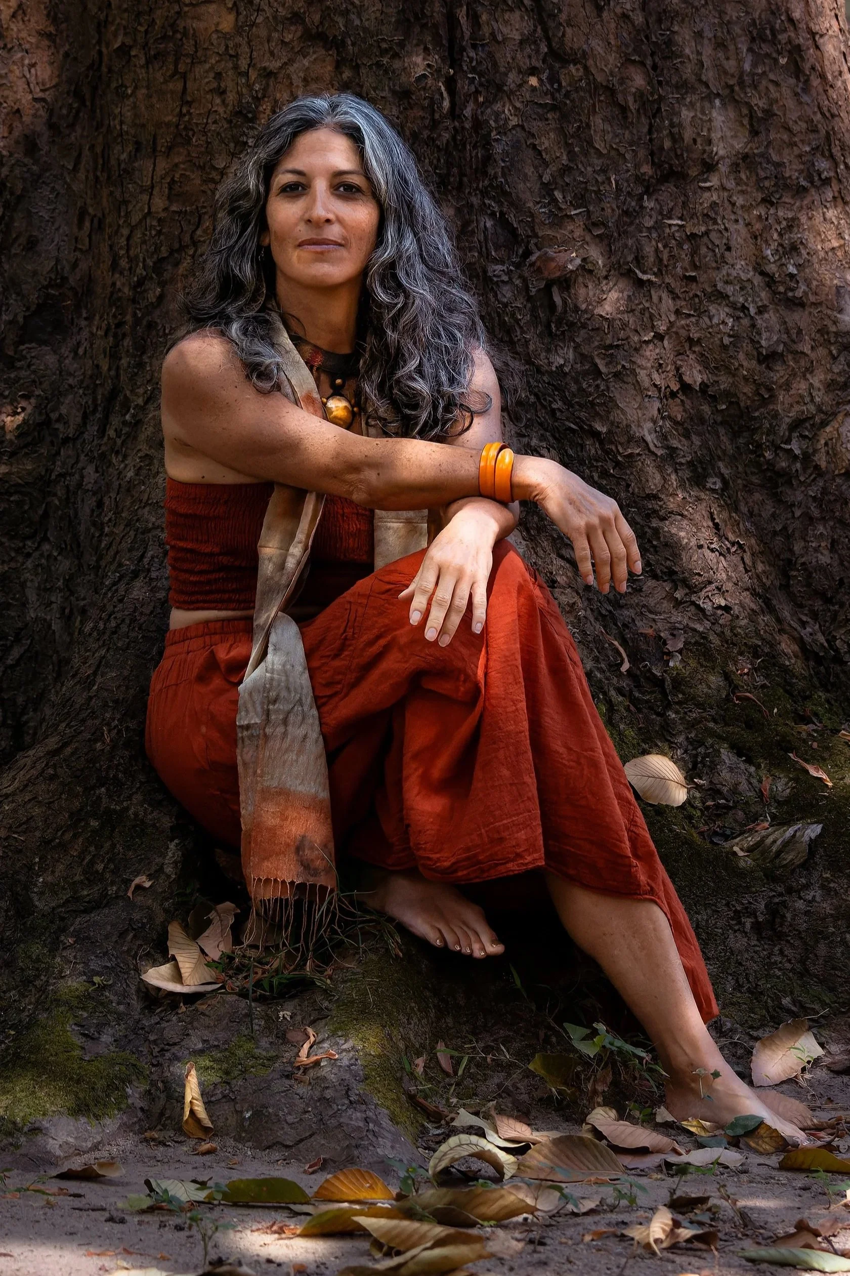A woman with long, wavy gray hair sitting against a large tree on the ground, surrounded by fallen leaves, dressed in an orange skirt and a beige shawl, with a large orange bracelet on her wrist.