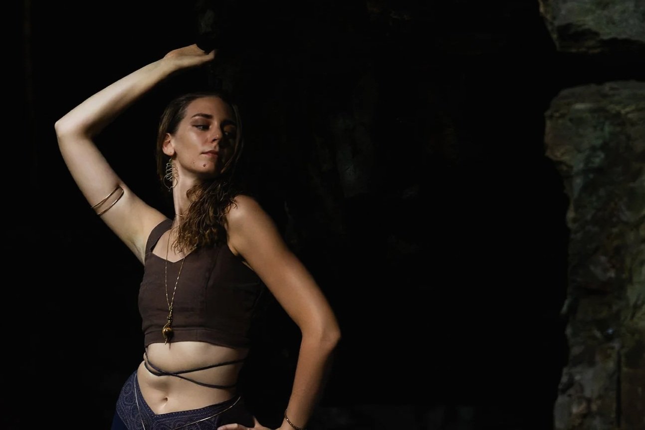 Young woman with long hair posing in a dark setting, wearing a cropped brown top, jewelry, and patterned pants.