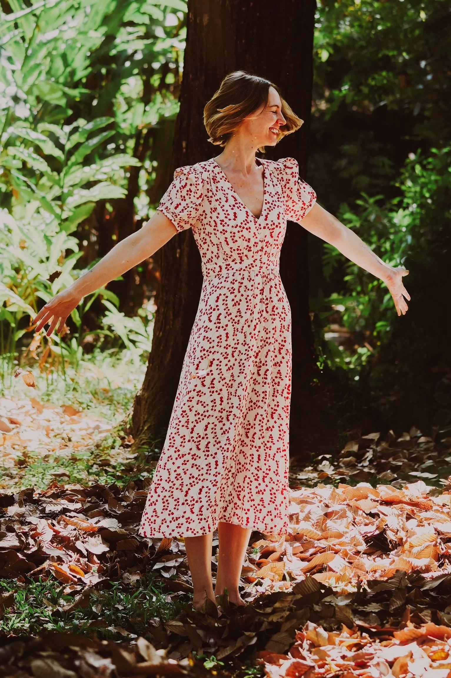 A woman in a long, white dress with red pattern, standing outdoors among trees and fallen autumn leaves, with arms outstretched and a joyful expression.