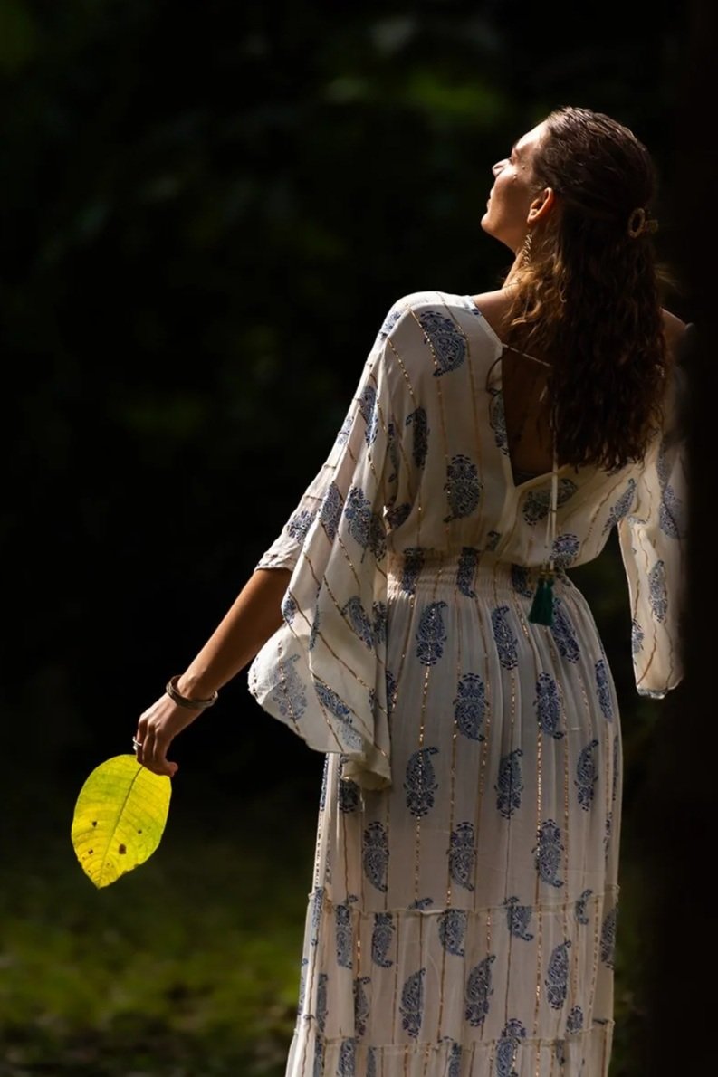 A woman wearing a patterned dress, holding a yellow leaf, standing outdoors in a dark, natural setting with her head tilted back and eyes closed.