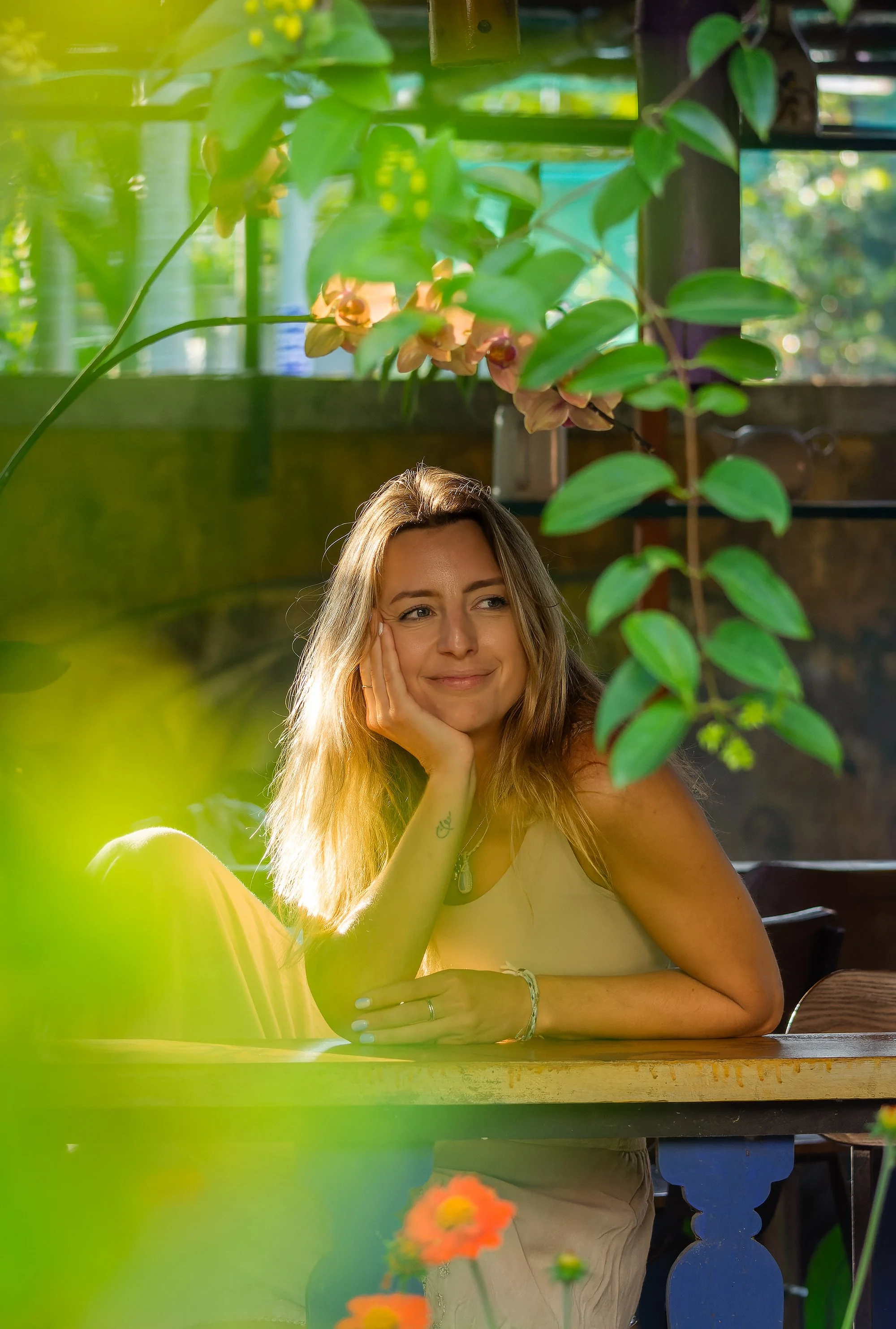 A young woman with long light brown hair sits at a wooden table in a lush, sunlit environment. She rests her head on her hand and smiles softly. Green plants and flowers surround her, with some flowers in the foreground and leaves hanging down.
