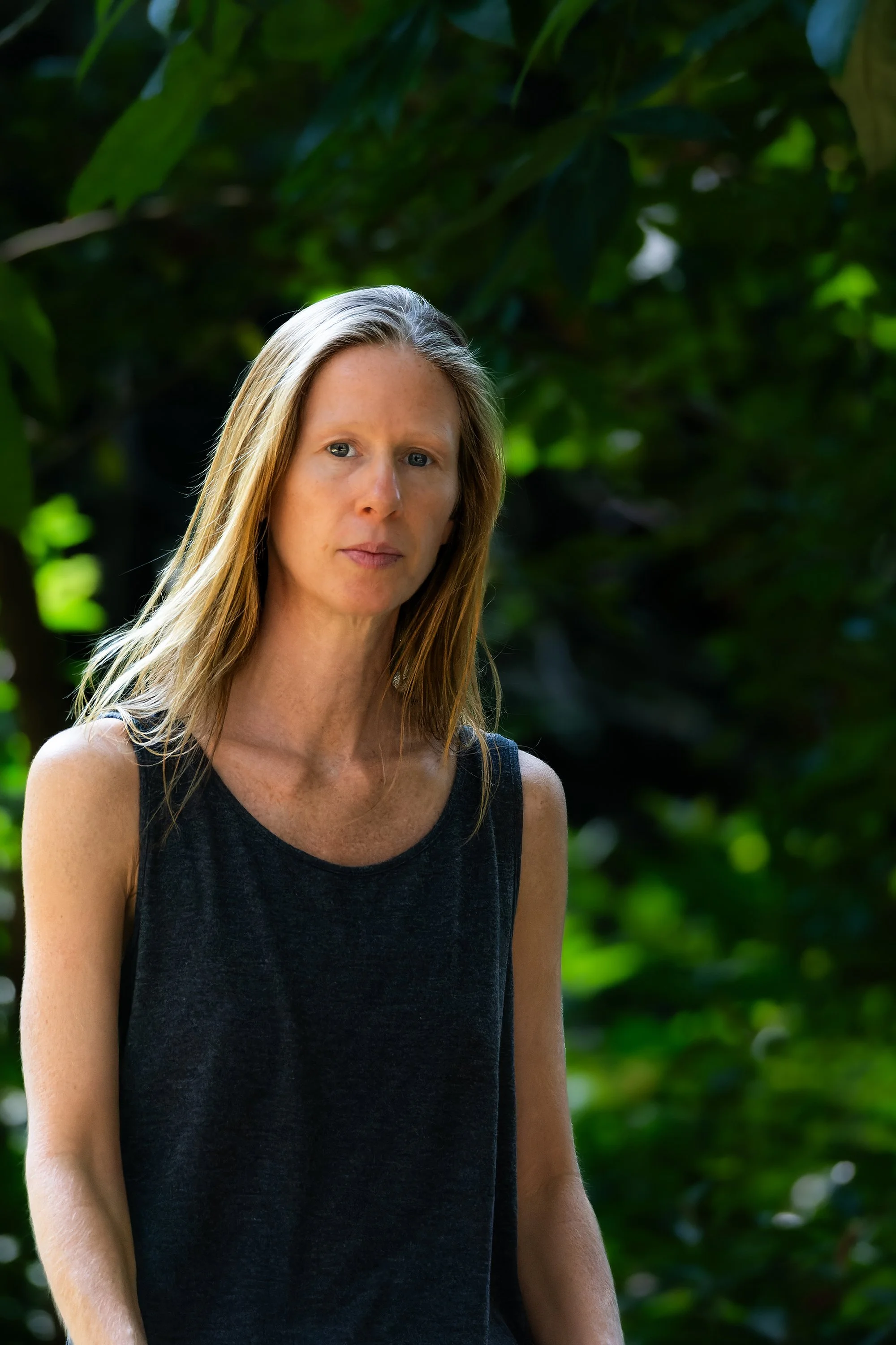 A woman with light skin and long blonde hair, wearing a black sleeveless top, standing outdoors with green foliage in the background.