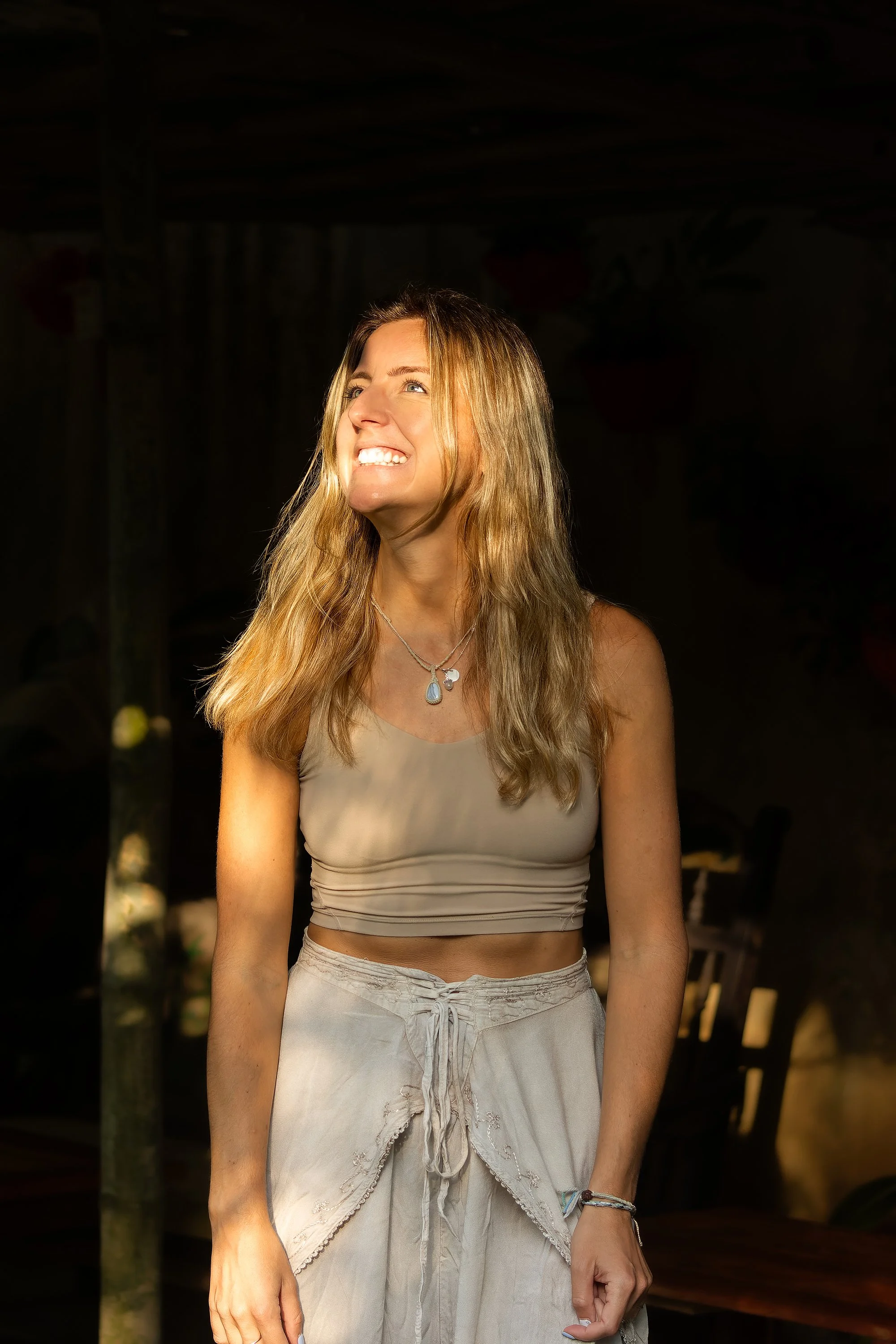 A young woman with long blonde hair, wearing a beige crop top and light-colored drawstring pants, smiling and looking to her left, standing in a dimly lit outdoor setting.