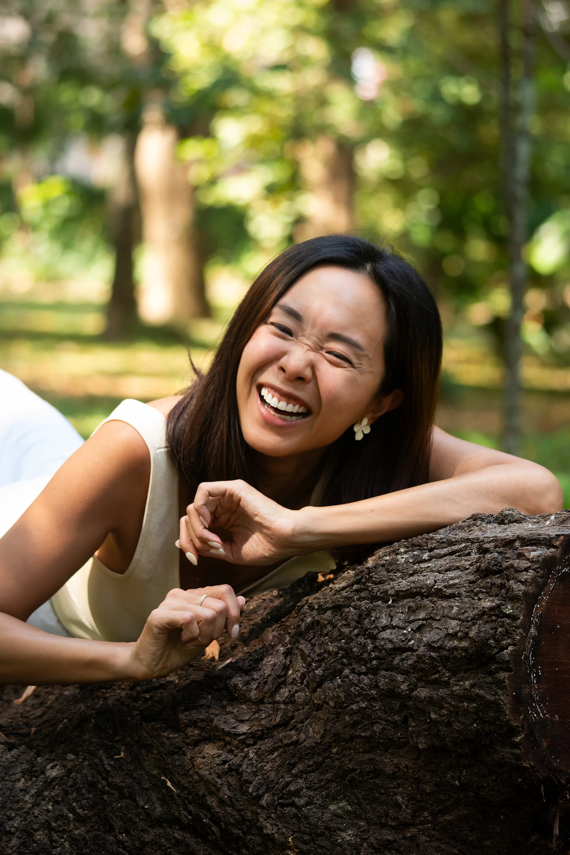 A woman is laughing and leaning on a fallen tree trunk in a park with green trees in the background.