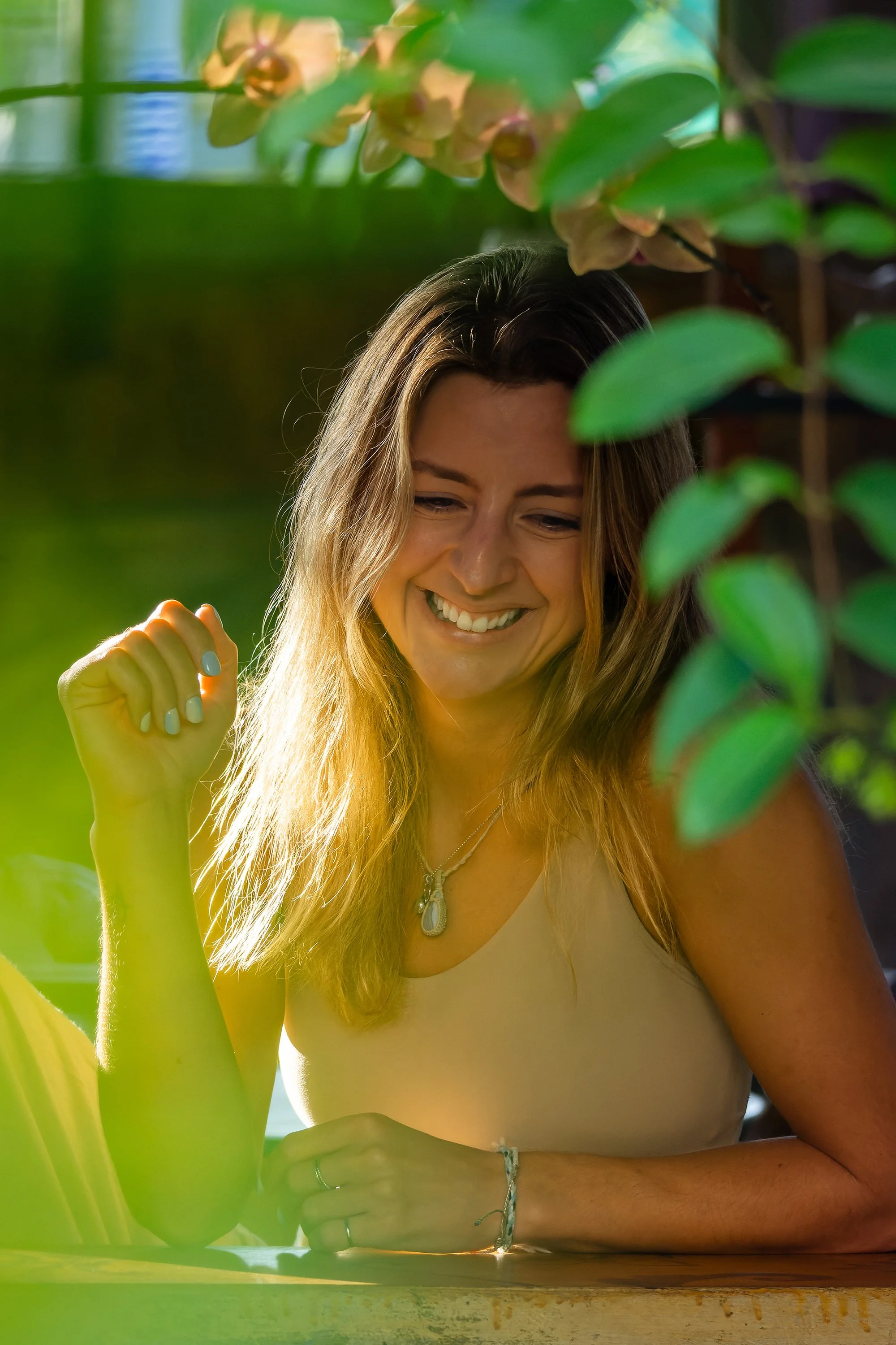A woman with long hair smiling, wearing a cream top and jewelry, sitting outdoors with green foliage around her.