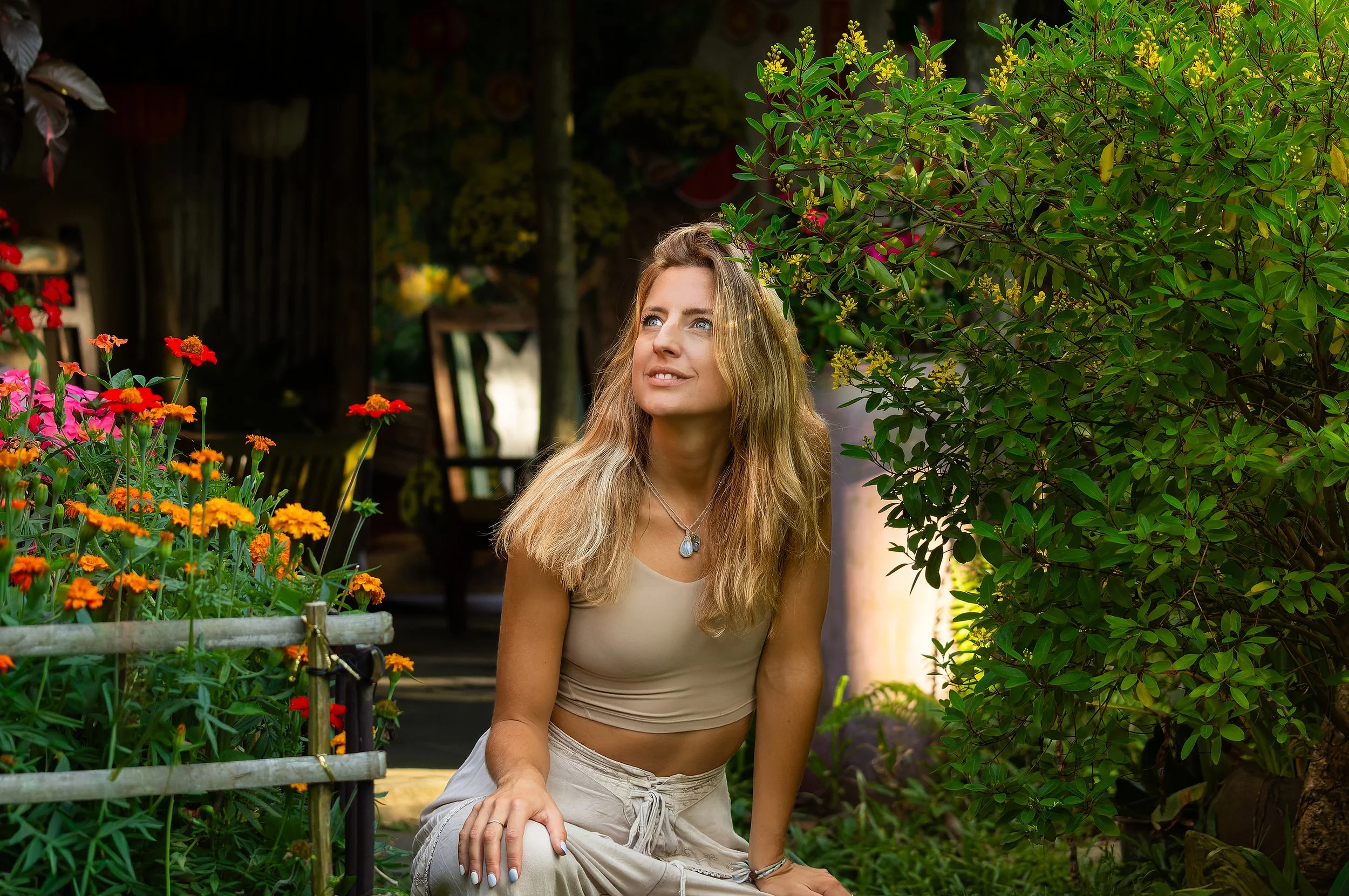 A young woman with long blonde hair and wearing a beige sleeveless top and light-colored pants, sitting in a garden among colorful flowers and green foliage, smiling and looking upward.