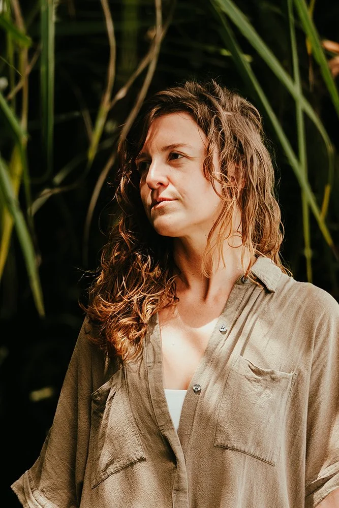 A woman with curly brown hair wearing a beige button-up shirt, standing outdoors among tall green plants.