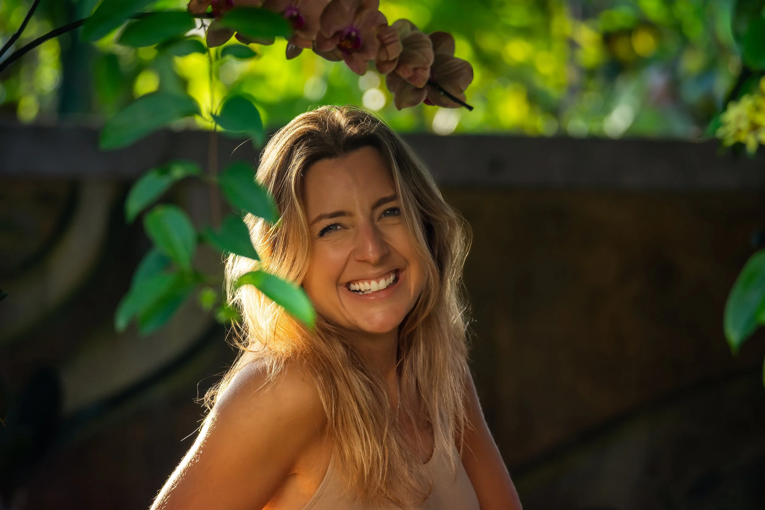 A smiling woman with long, light brown hair outdoors, partially obscured by green leaves and pink flowers with blurred green foliage and sunlight in the background.