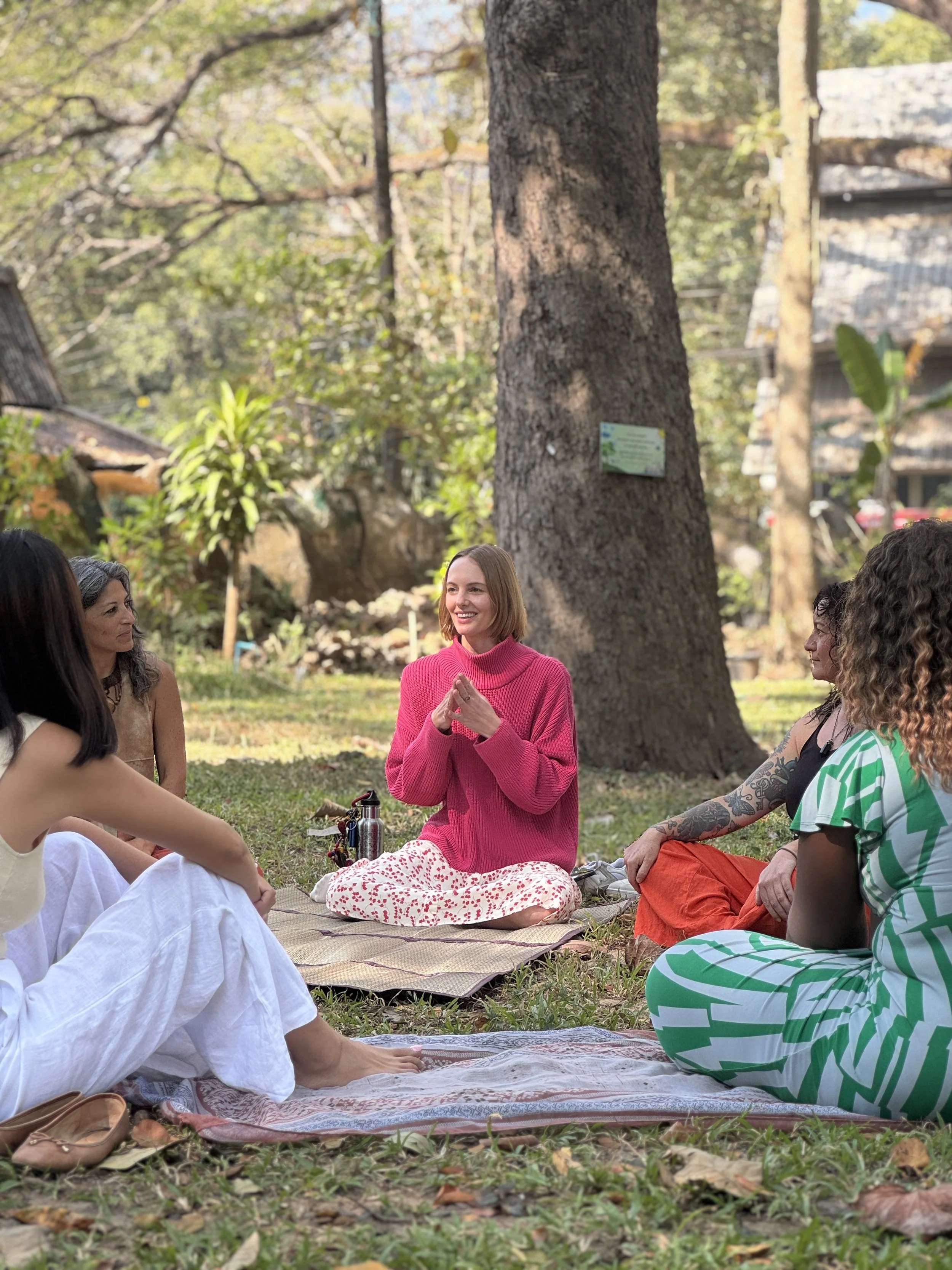 A group of diverse people sitting on a blanket on the grass in a park, engaged in a meditation or discussion. There is a large tree in the background and autumn leaves scattered on the ground. The group includes women dressed in comfortable, colorful clothing, with one woman in a bright pink sweater and patterned pants smiling and clasping her hands.