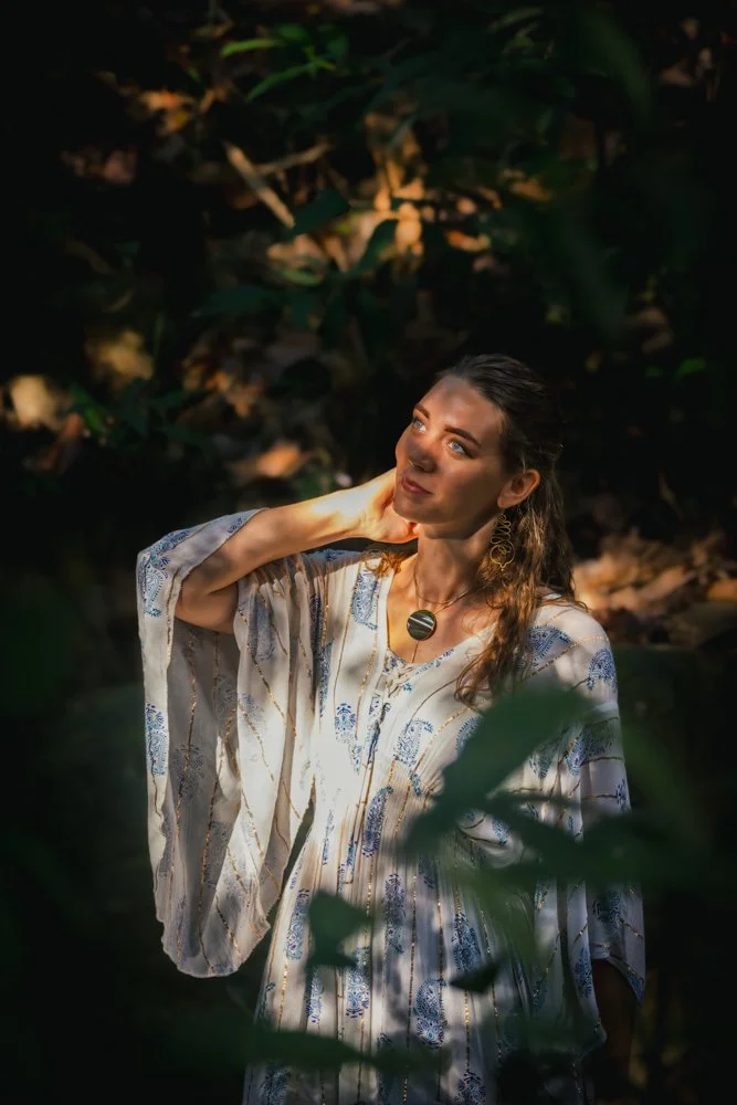 A woman with long brown hair and blue eyes, wearing a patterned flowy dress and large earrings, standing in a lush green forest with sunlight filtering through the leaves.