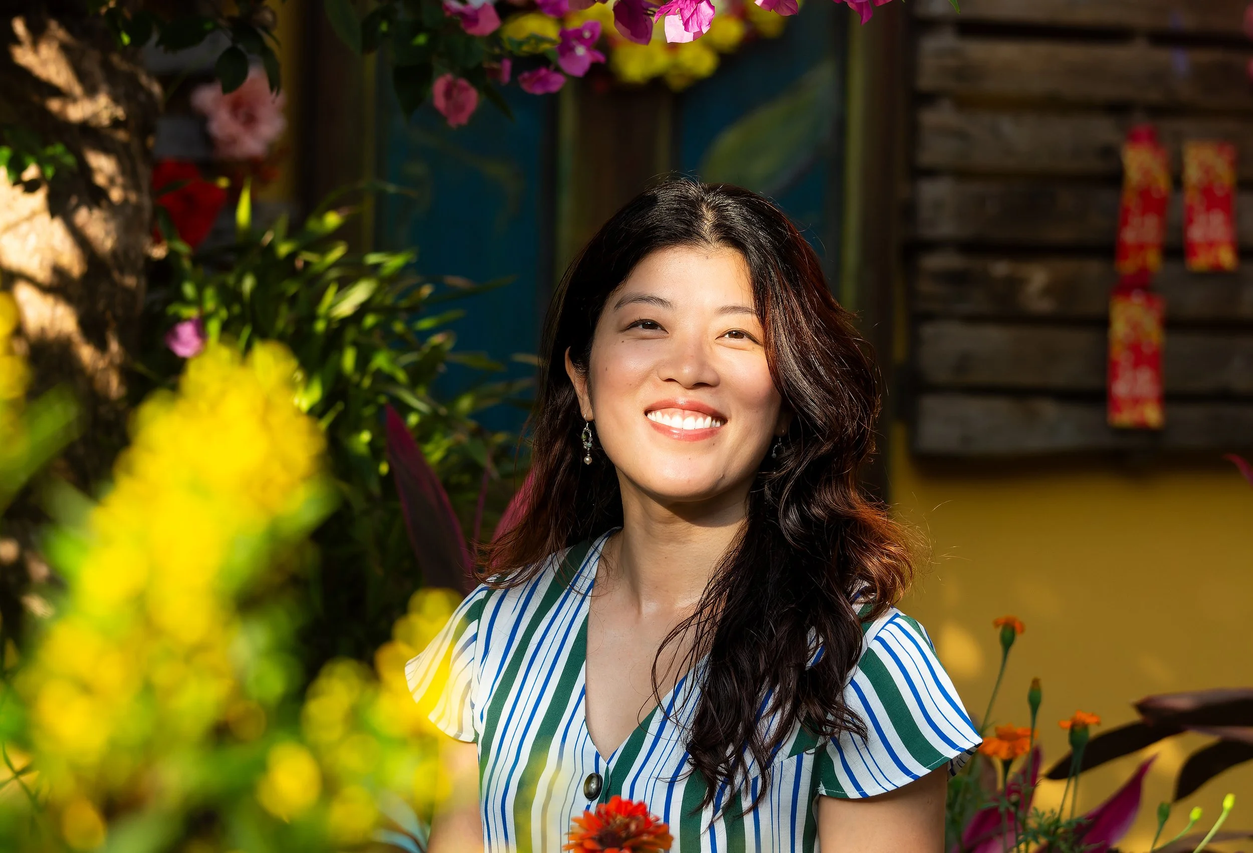 A smiling woman with long, dark, wavy hair and earrings, wearing a striped shirt, is standing outdoors among colorful flowers and plants, with a wooden wall and decorations in the background.