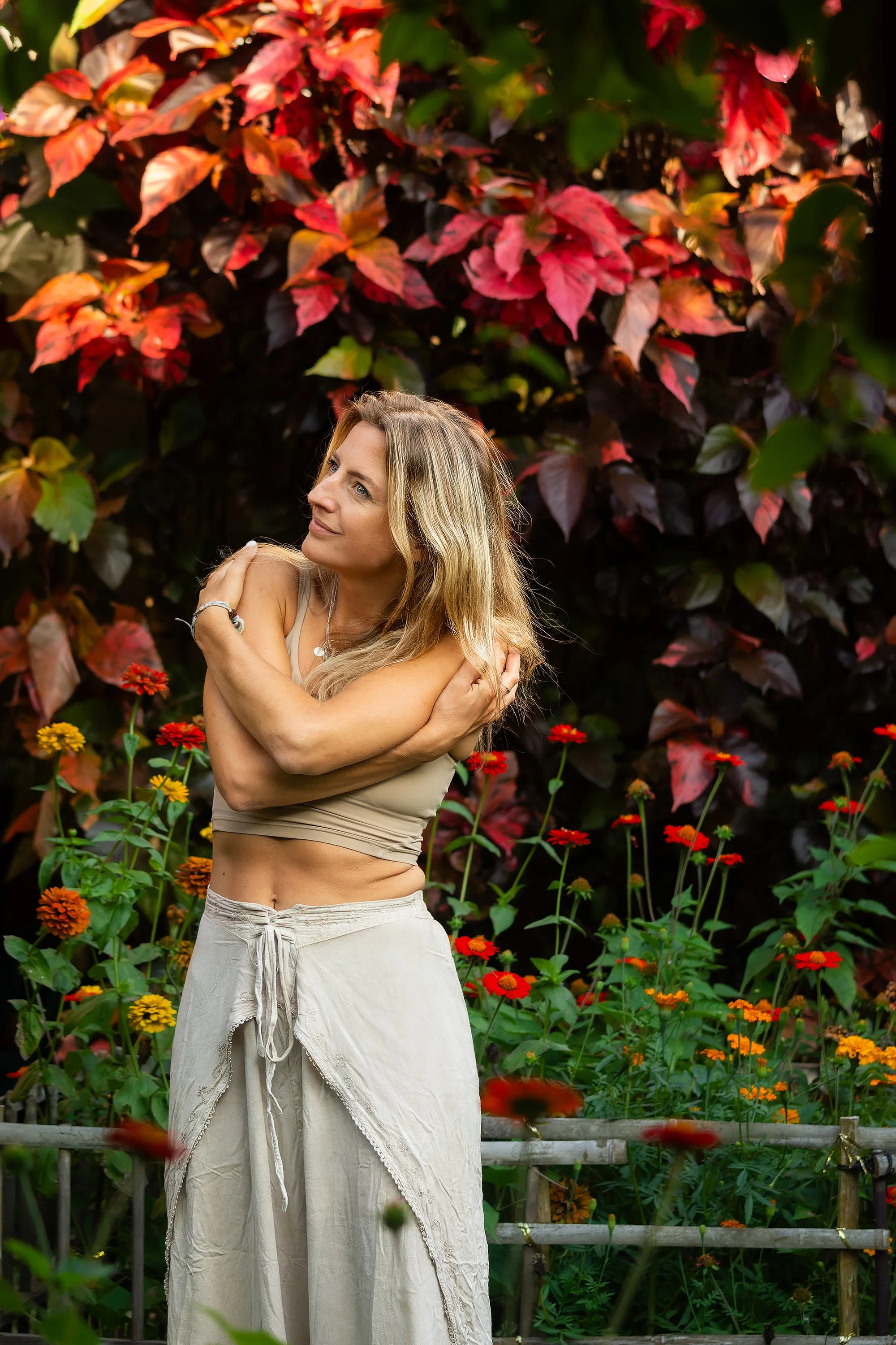 A woman with long blonde hair wearing a beige crop top and loose beige pants standing outdoors amidst red and orange flowers and dark green leaves.